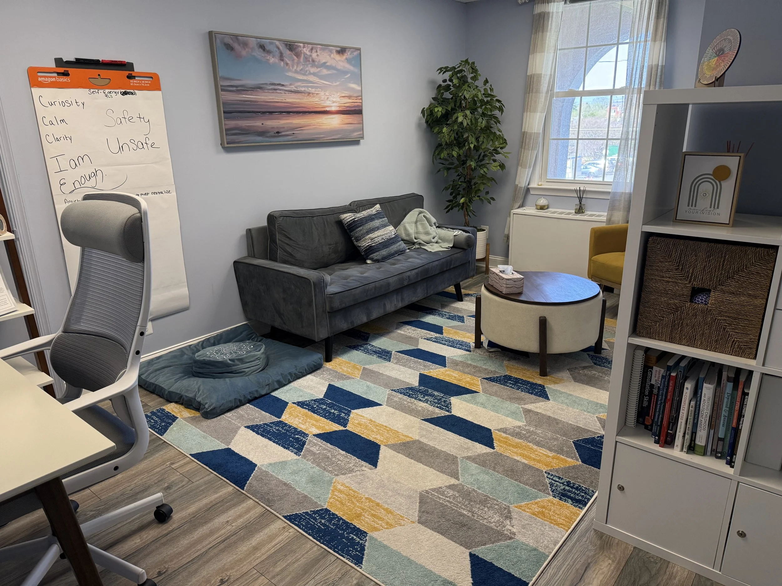Living room with a gray couch, a colorful geometric rug, and a large window with sheer curtains, decorated with framed artwork, bookshelves, and a yellow chair.