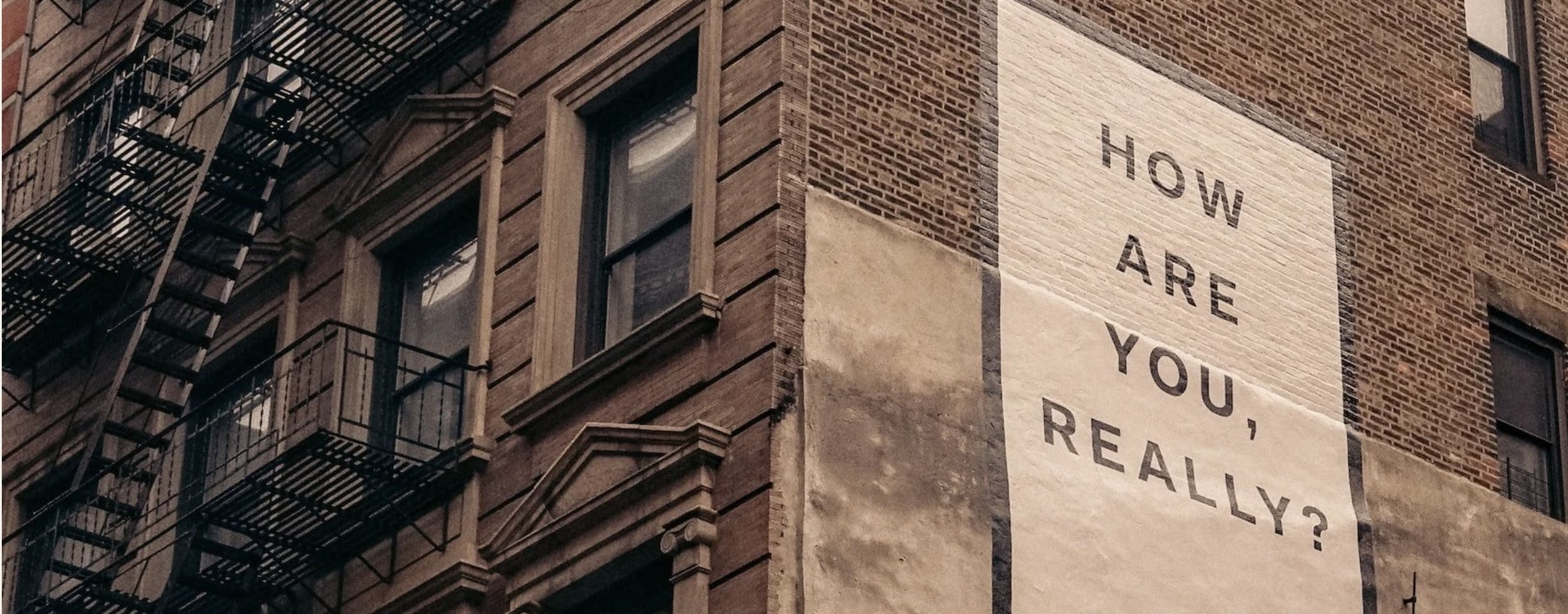 A brick building in Brooklyn with multiple windows and a black fire escape. On the side of the building, there is a large white banner with the words 'HOW ARE YOU, REALLY?' written in black letters.