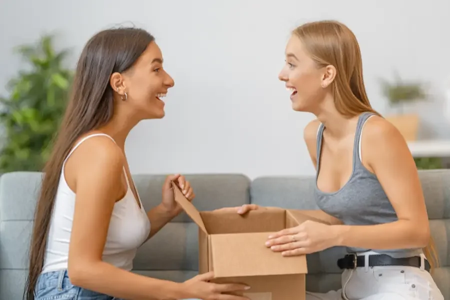 Two young women sit on a couch, smiling and holding a box between them, suggesting a moment of sharing or gift-giving.