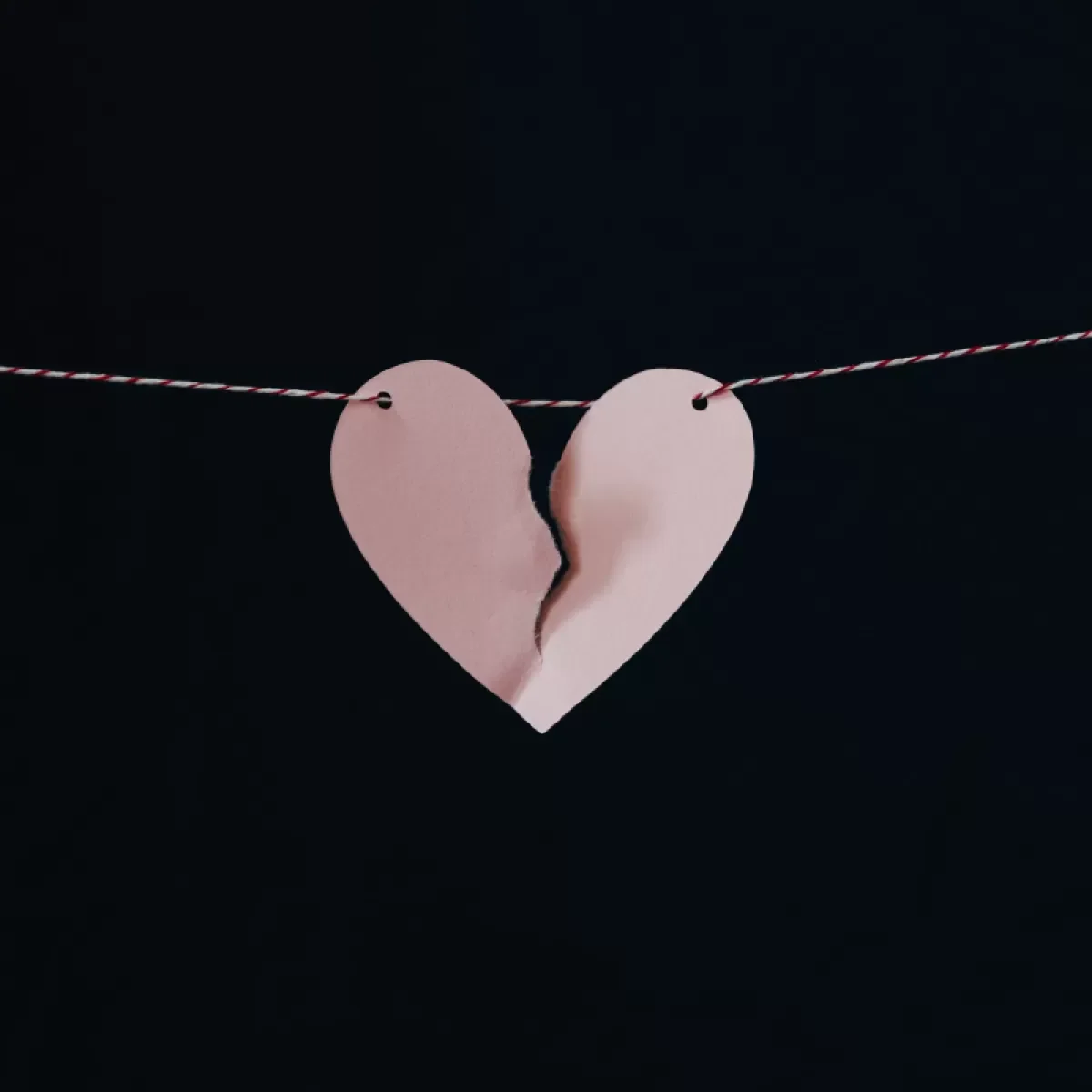 Pink paper heart with a jagged tear hanging from a red and white striped string against a black background.