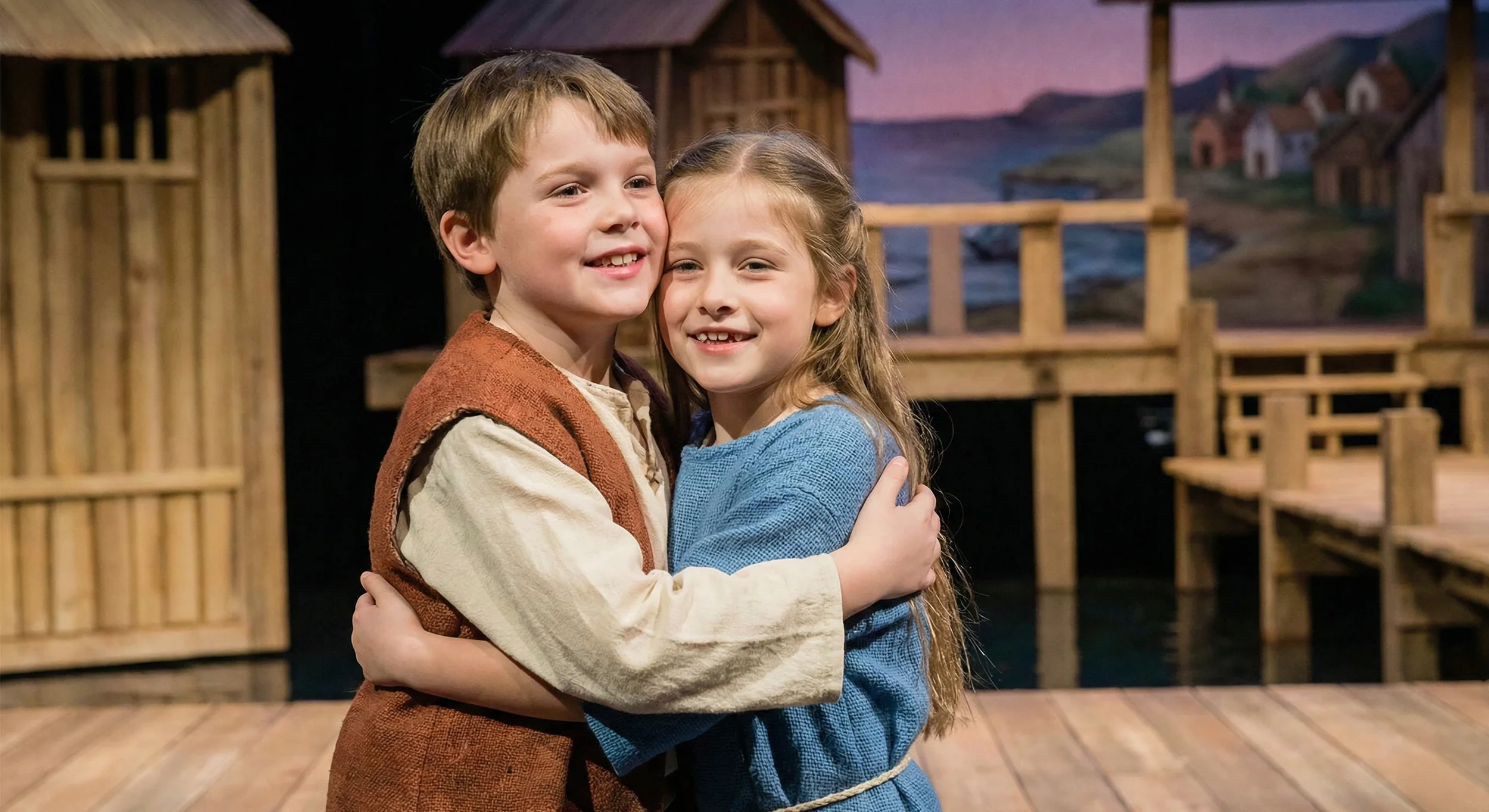 Two children, a boy and a girl, hugging and smiling on a wooden stage with a backdrop depicting a village scene with houses and a river.