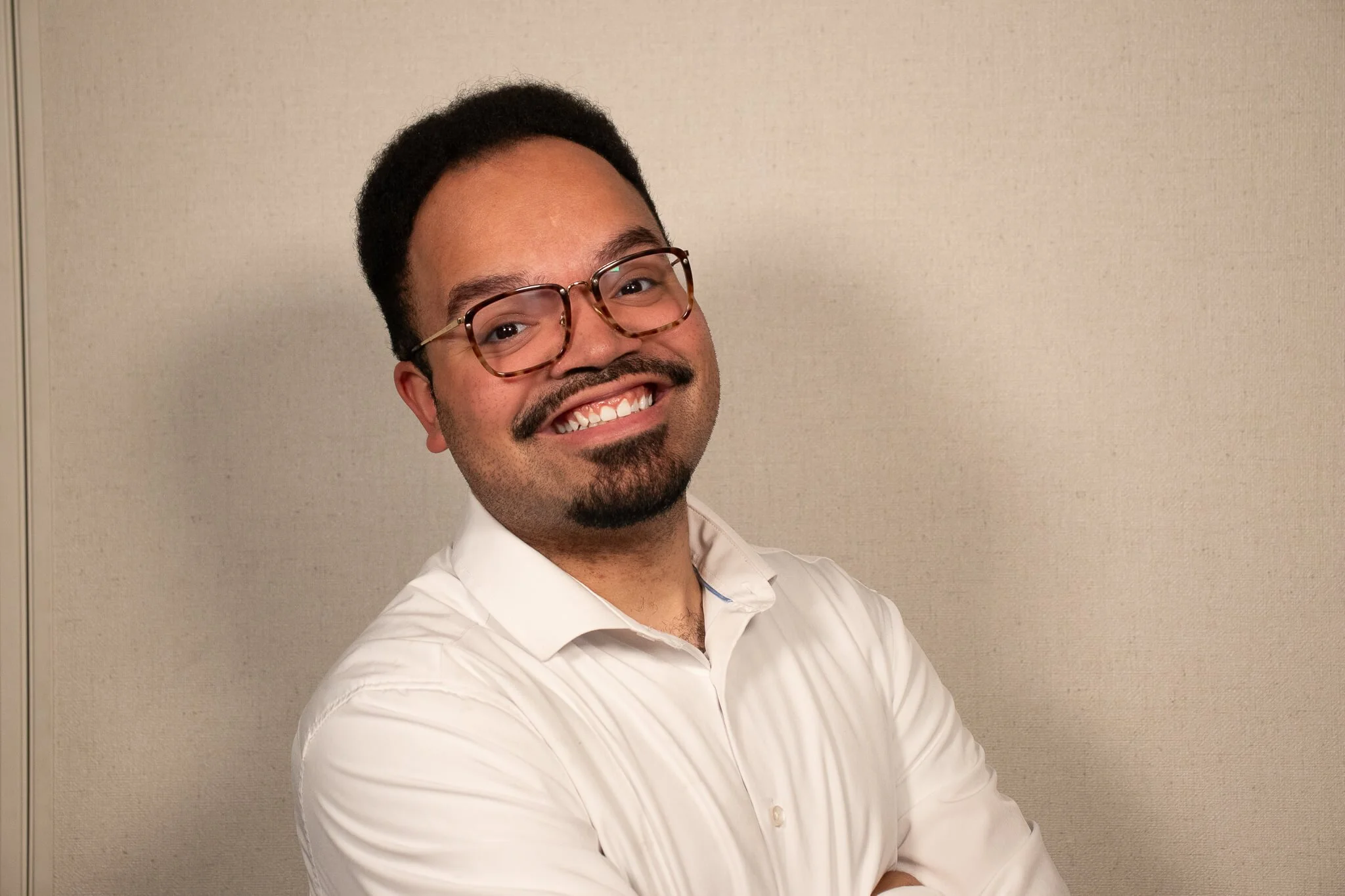 A man in a white shirt, with black hair and glasses smiling in front of a tan back drop.