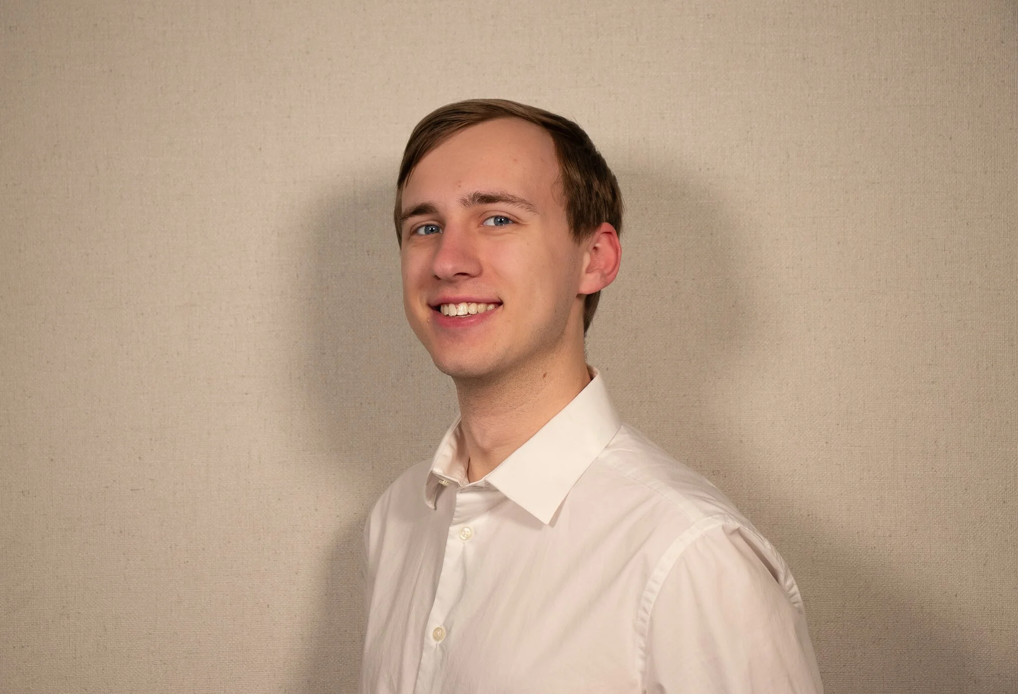 A young man with light brown hair and blue eyes, wearing a white shirt, smiling against a plain tan background.