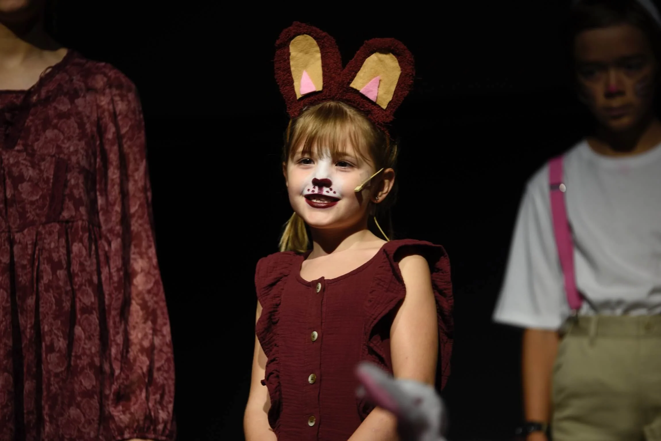 A young girl wearing a maroon dress, felt animal ears, and face makeup smiles on stage during a performance.