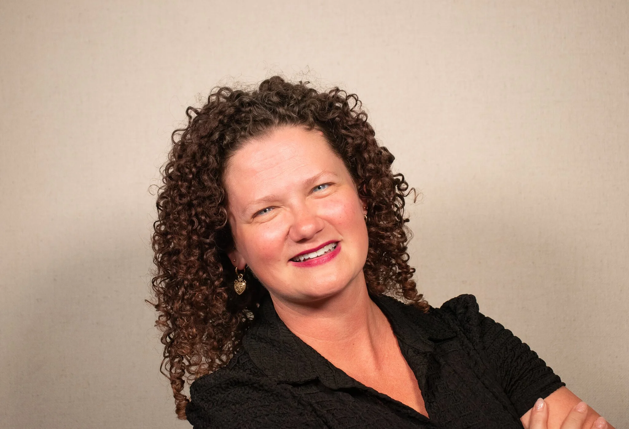 A woman with curly brown hair, blue eyes, and a black shirt, smiling in front of a tan background.