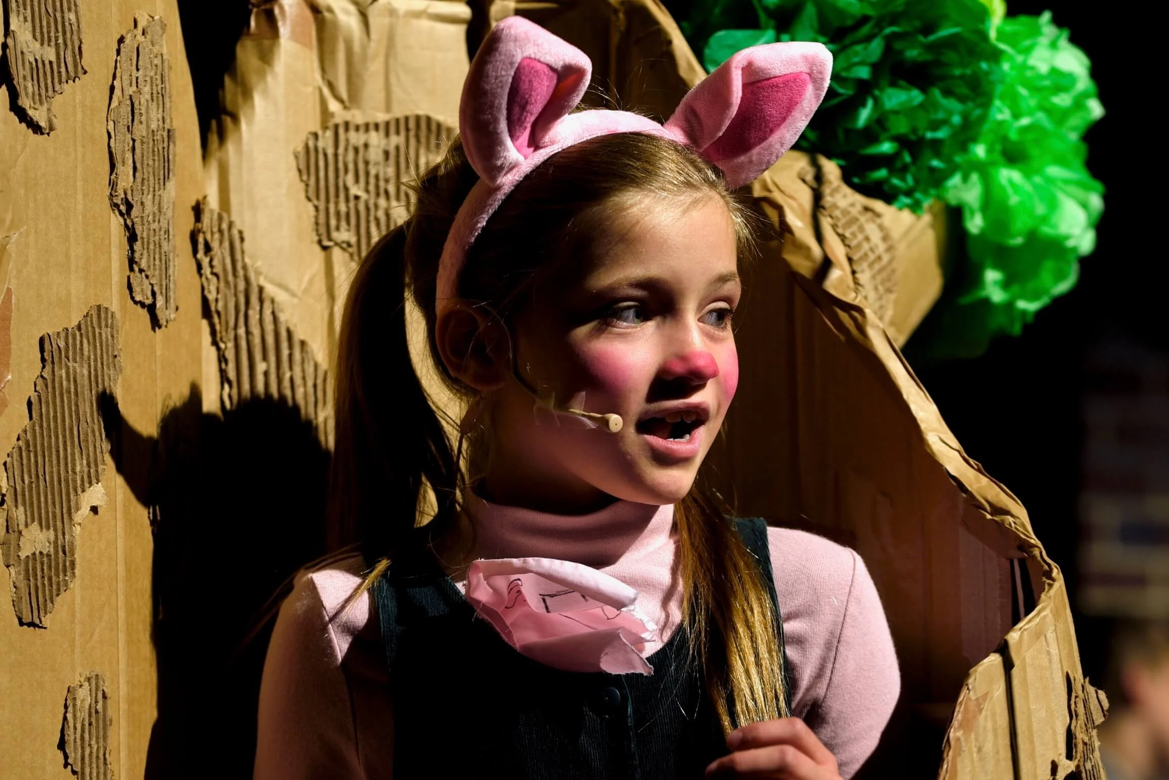 Close-up of a young girl playing Piglet in pink ears and nose makeup smiling and speaking on stage in front of a cardboard tree set piece.