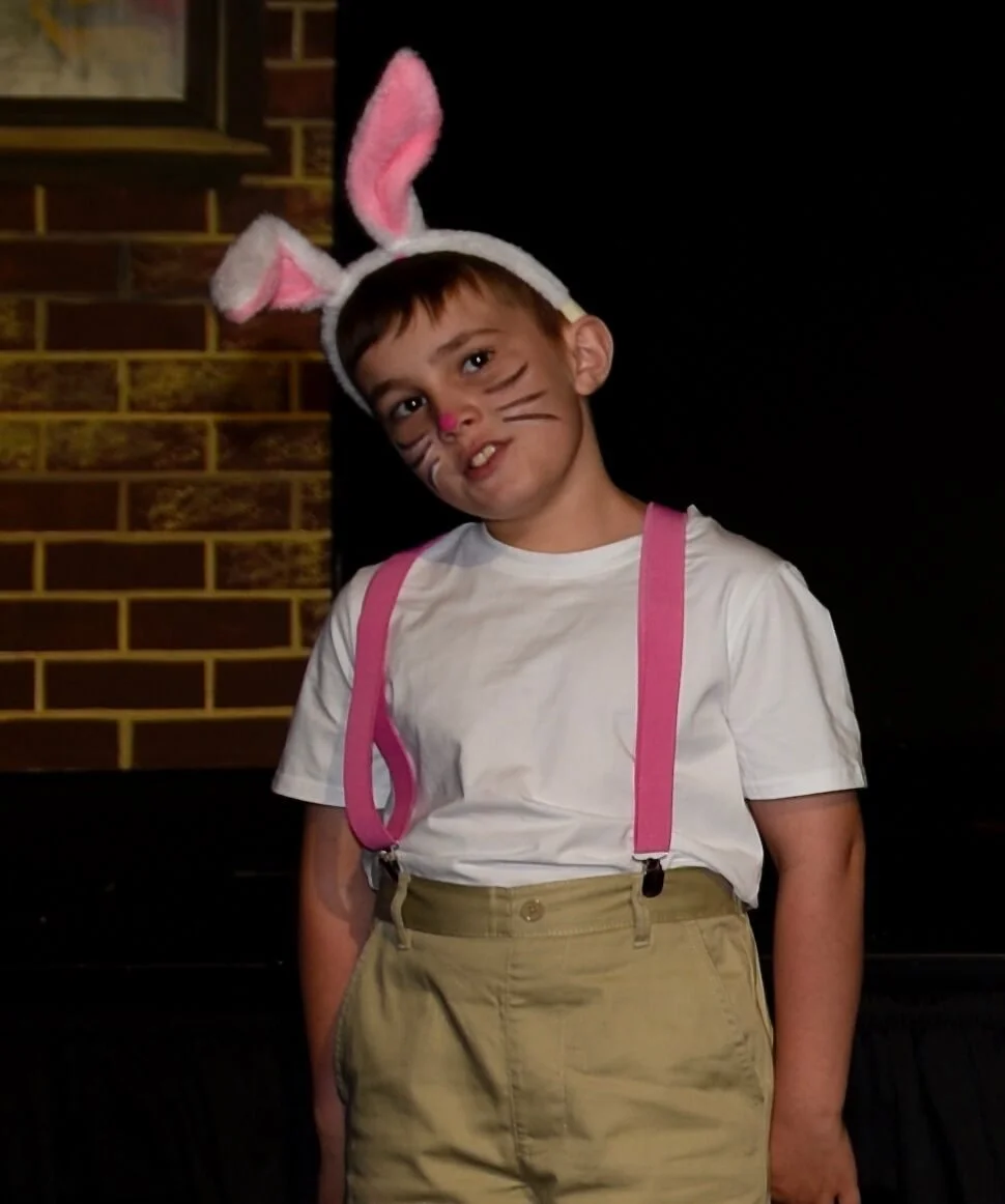 A young actor in rabbit ears, pink suspenders, and whisker makeup posing with his head tilted inquiringly on stage.