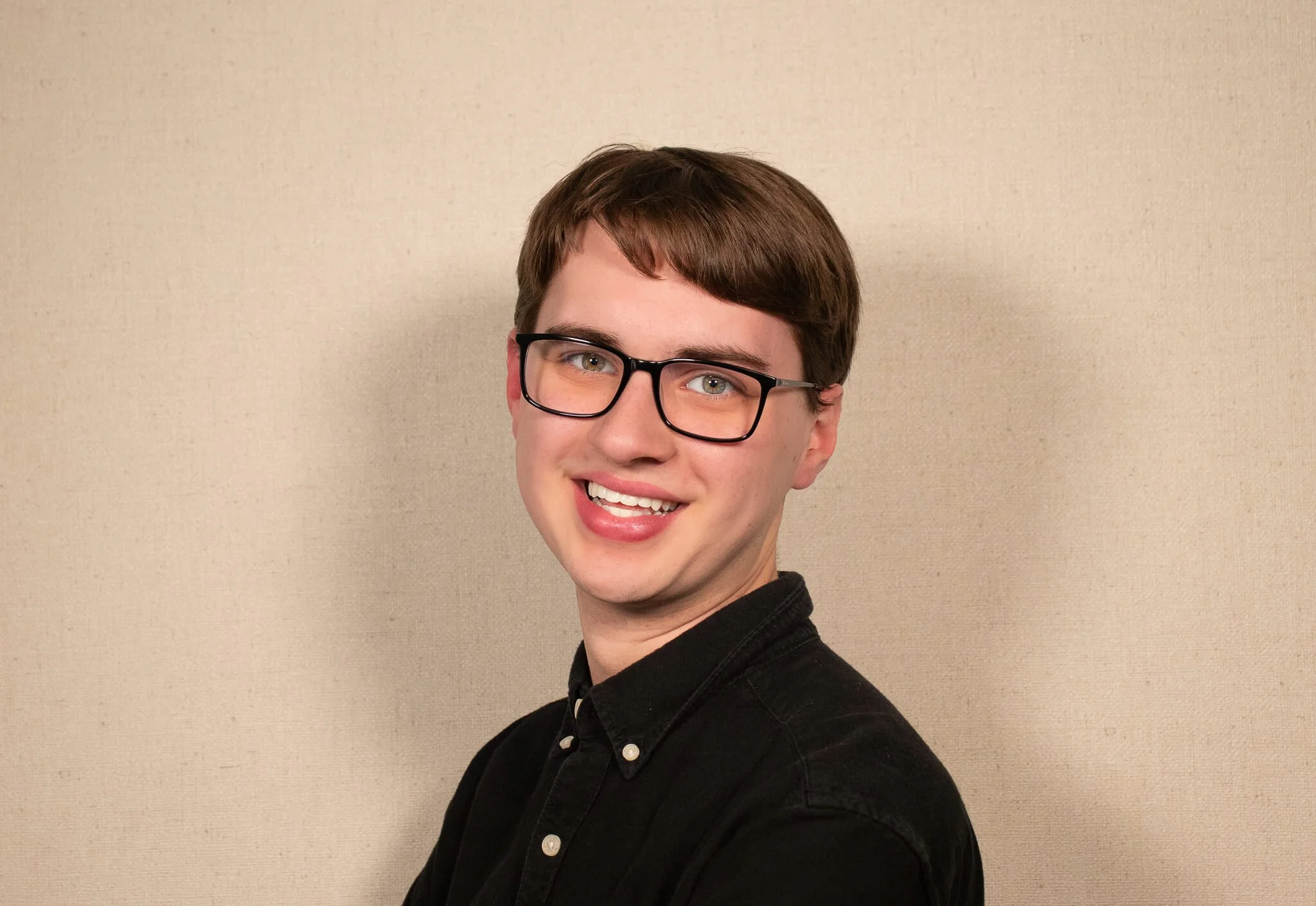 Portrait of a young man with short brown hair, glasses, smiling, wearing a black shirt against a tan background.