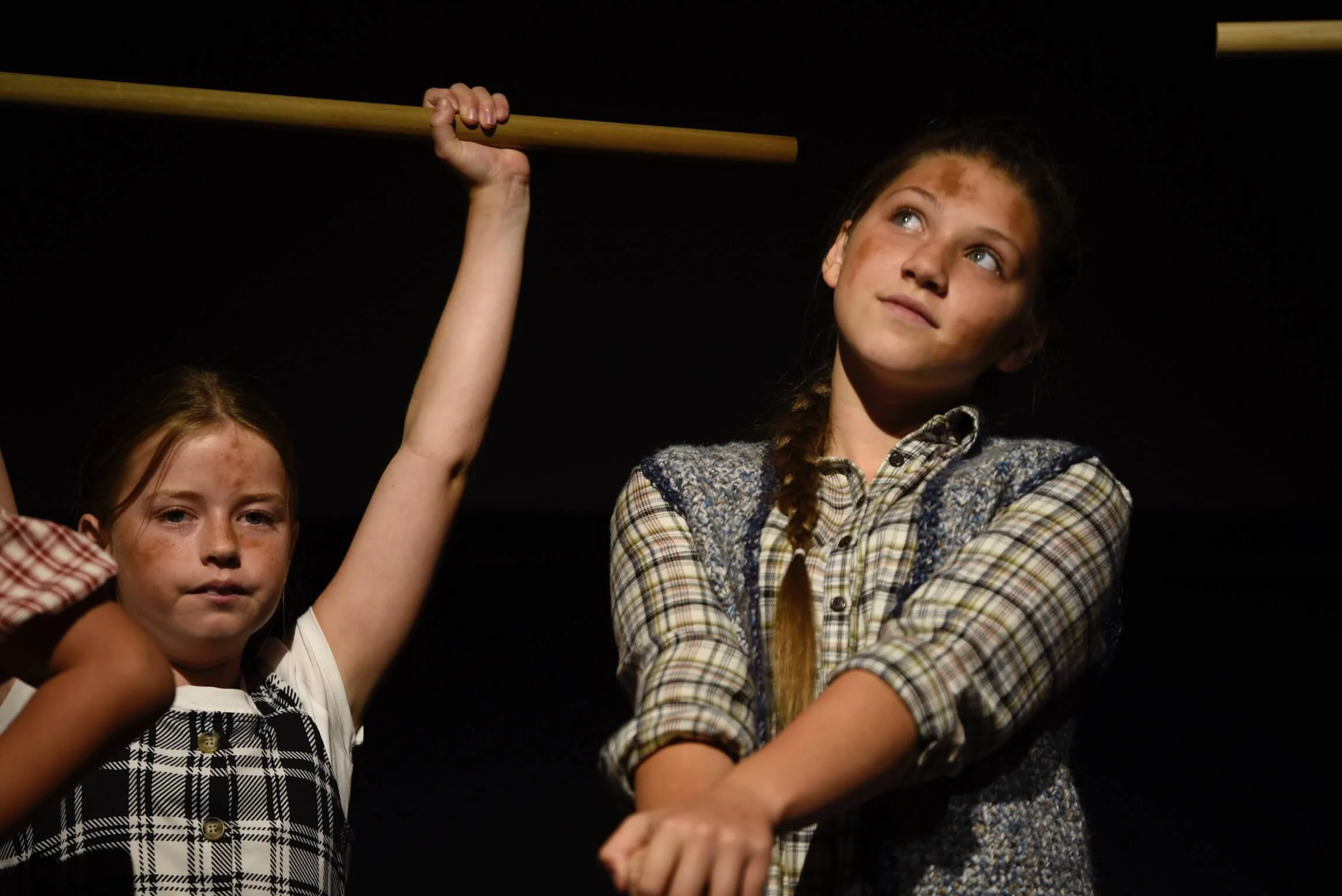 Two young actors with dirt-smudged faces holding wooden poles on stage, one looking upward thoughtfully.