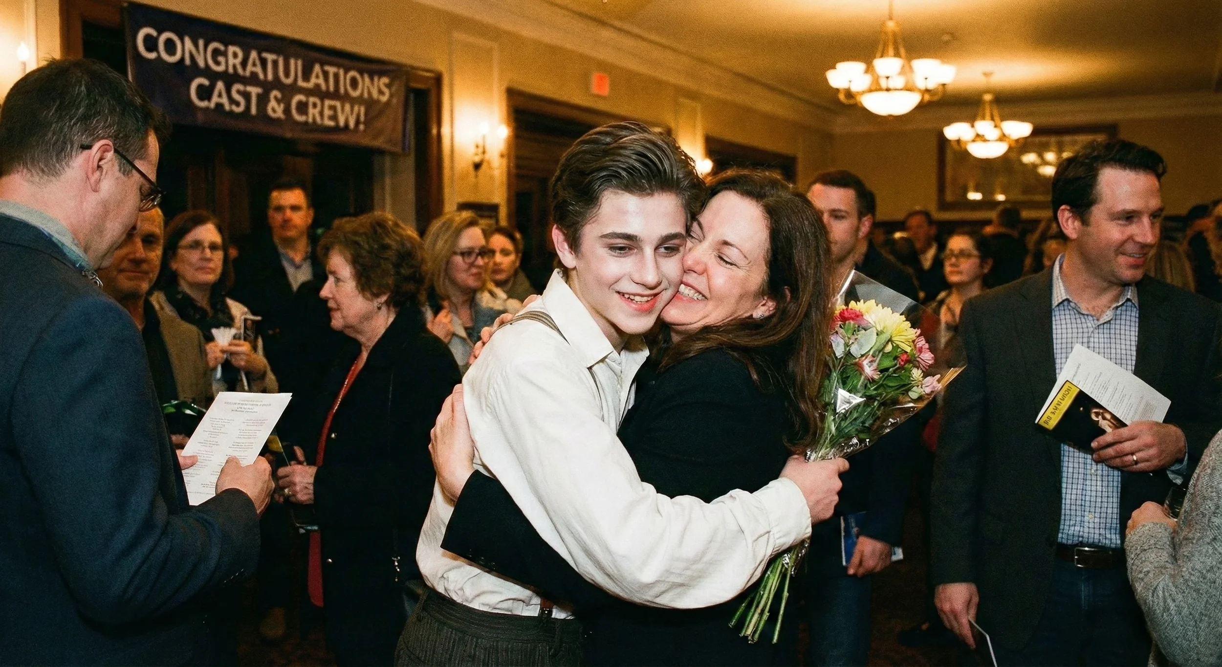 A young man and a woman hugging; the woman holds a bouquet of colorful flowers. In the background, there are other people at an event, with a banner that says 'Congratulations Cast & Crew!'