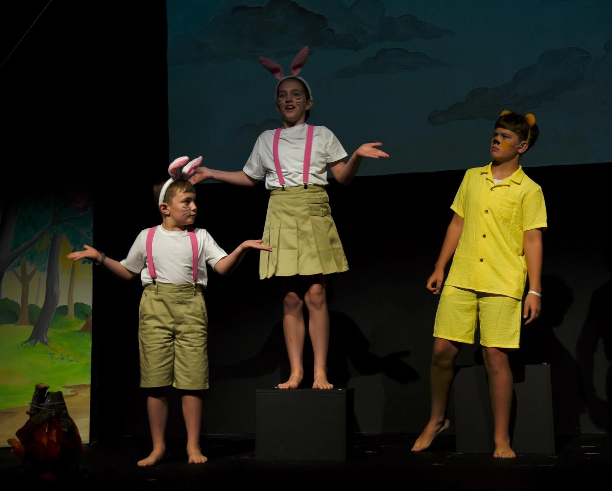 Three young actors on stage during a play: two dressed as rabbits gesturing with open hands while a third, dressed as Winnie the Pooh in yellow, looks on with a comical expressions.