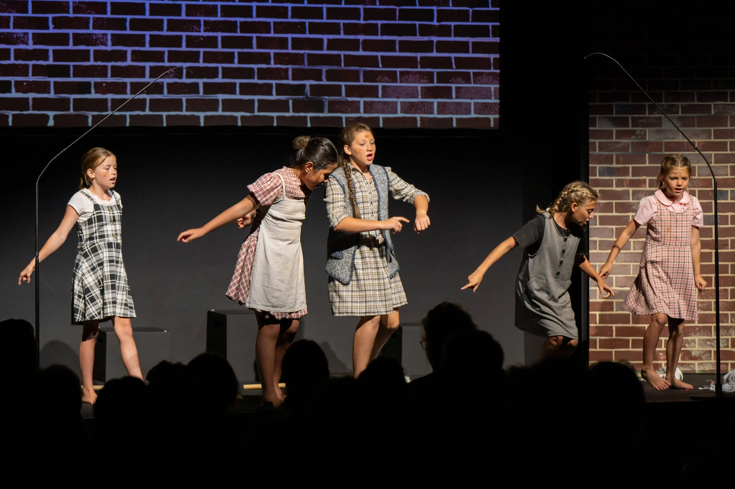 A group of young actors dressed as orphans singing and dancing in front of a brick wall backdrop.