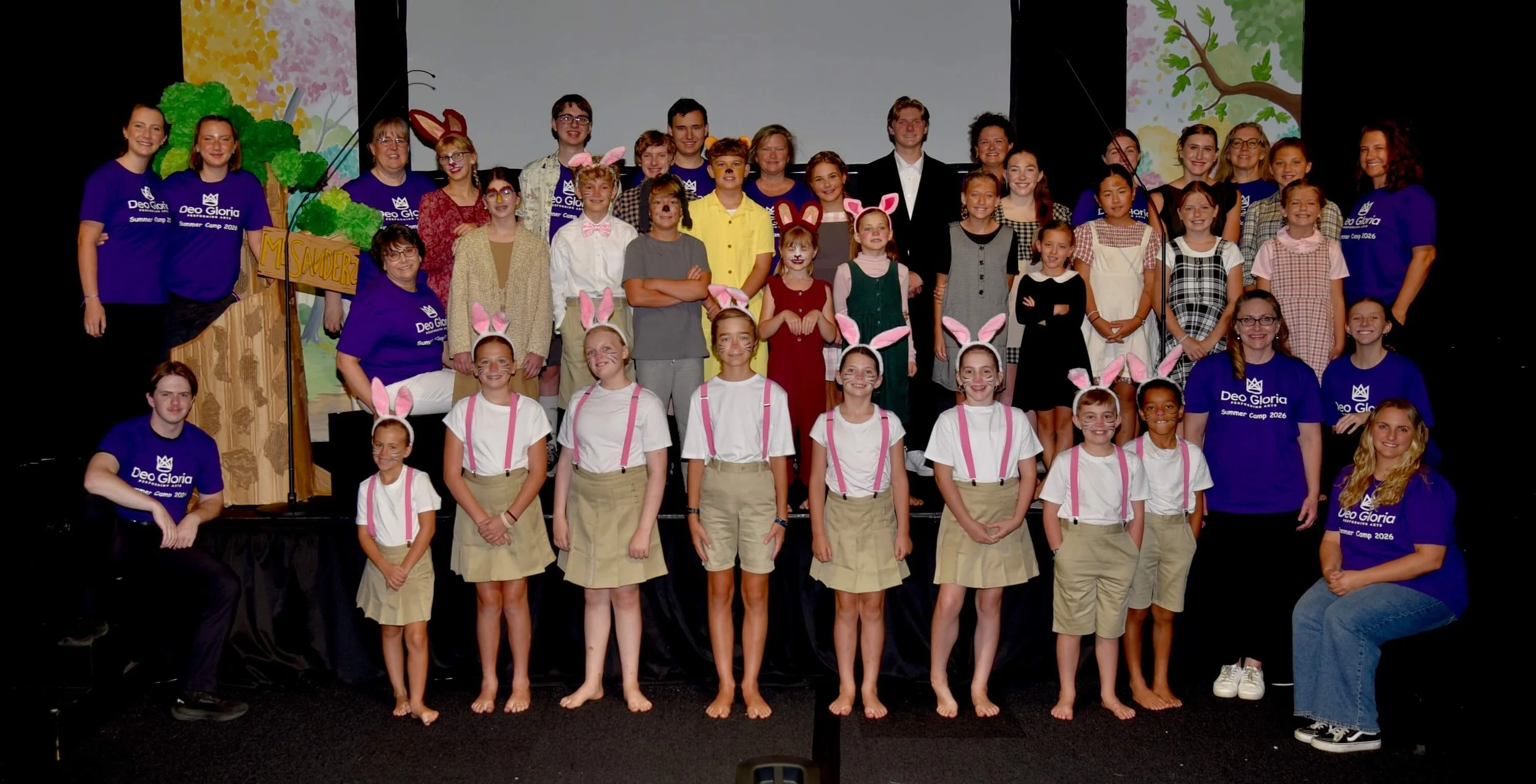 A large group photo on stage featuring the full cast and crew of a theater production, with costumed young performers standing alongside crew members wearing matching purple t-shirts.