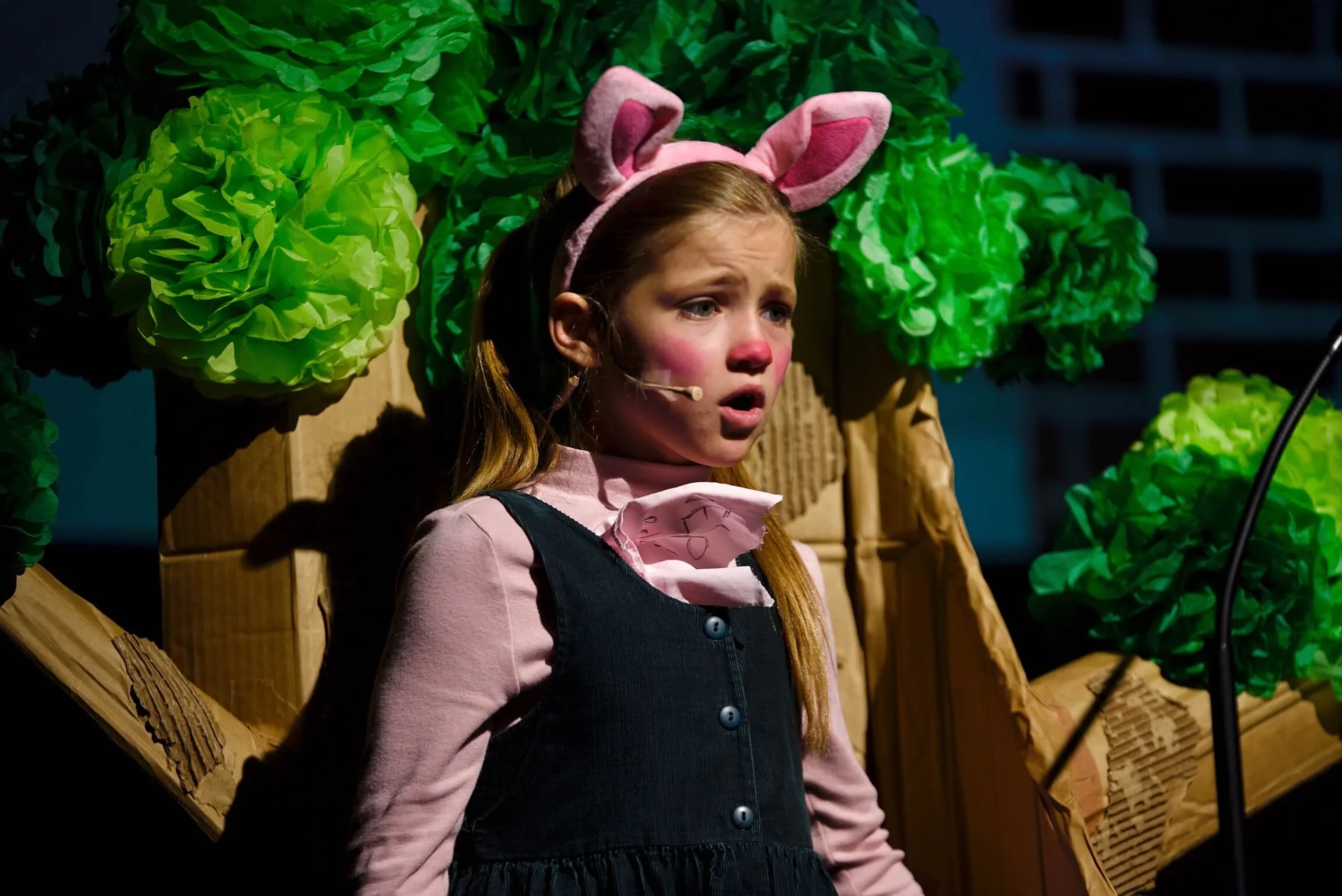 Close-up of a young girl playing Piglet in pink ears and nose makeup, looking startled on stage in front of a paper tree set piece.