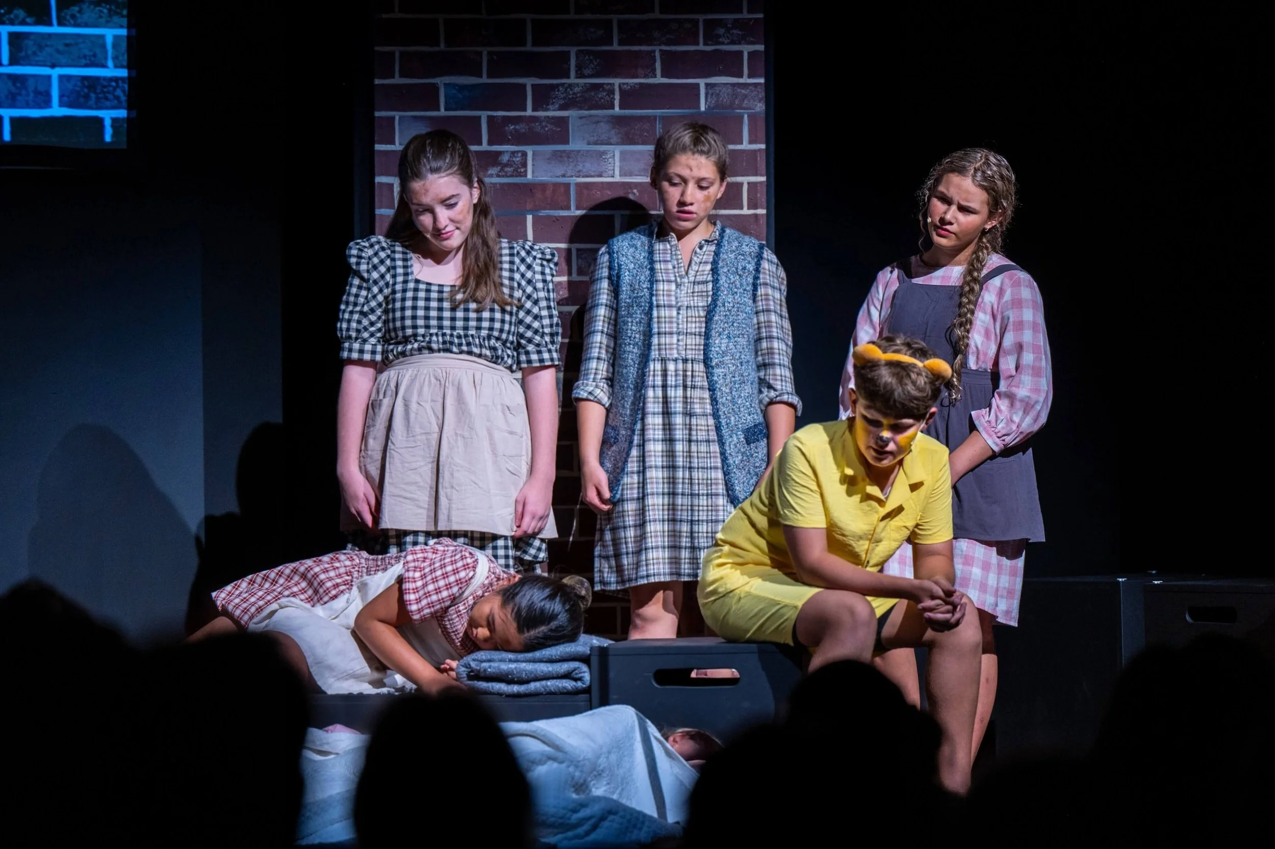 Five young actors wearing plaid and vintage attire look downcast during a stage performance against a brick backdrop.