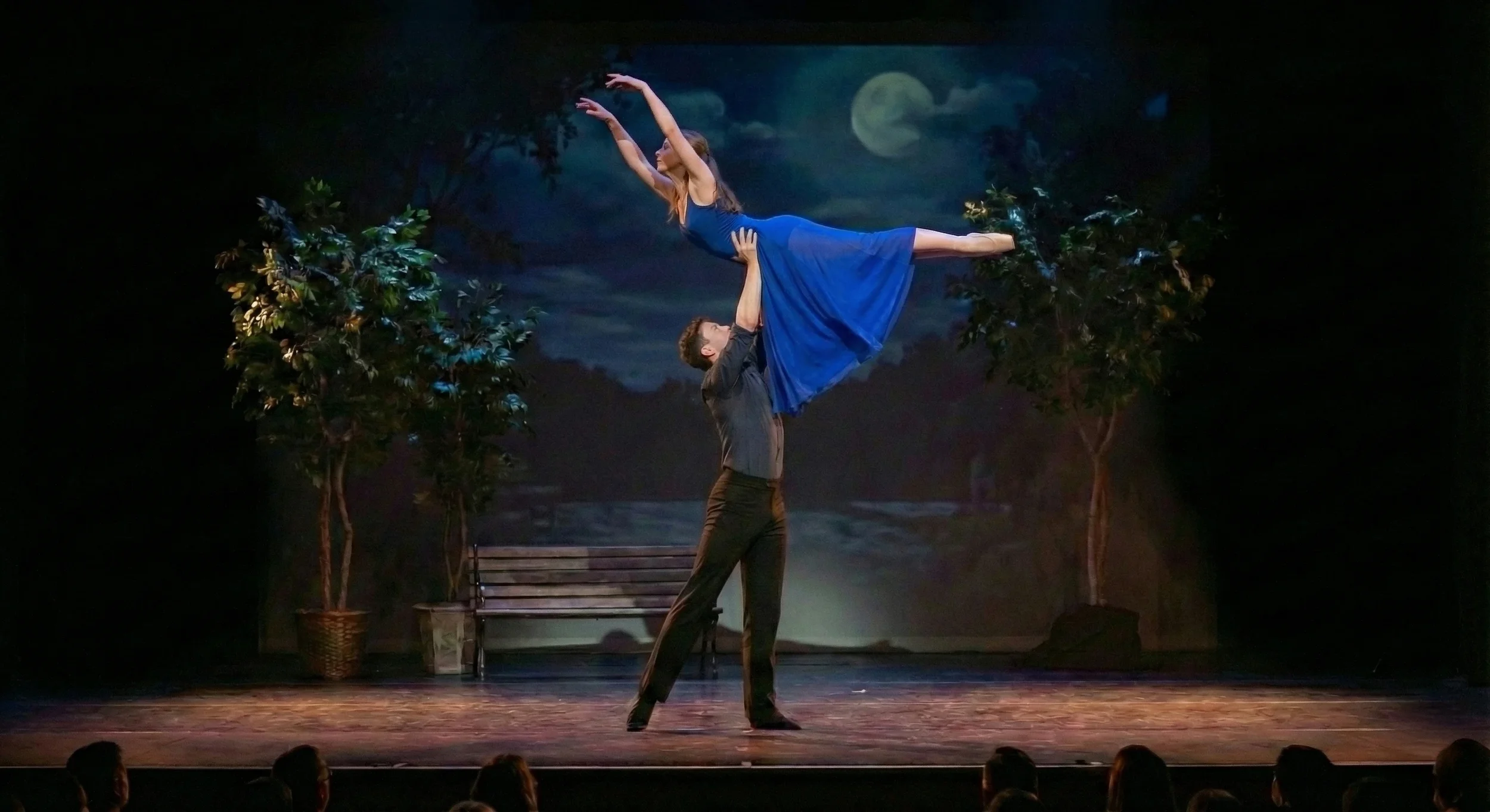 A ballet performance on stage with a male dancer lifting a female dancer in a blue dress, set against a backdrop of a moonlit night with trees.