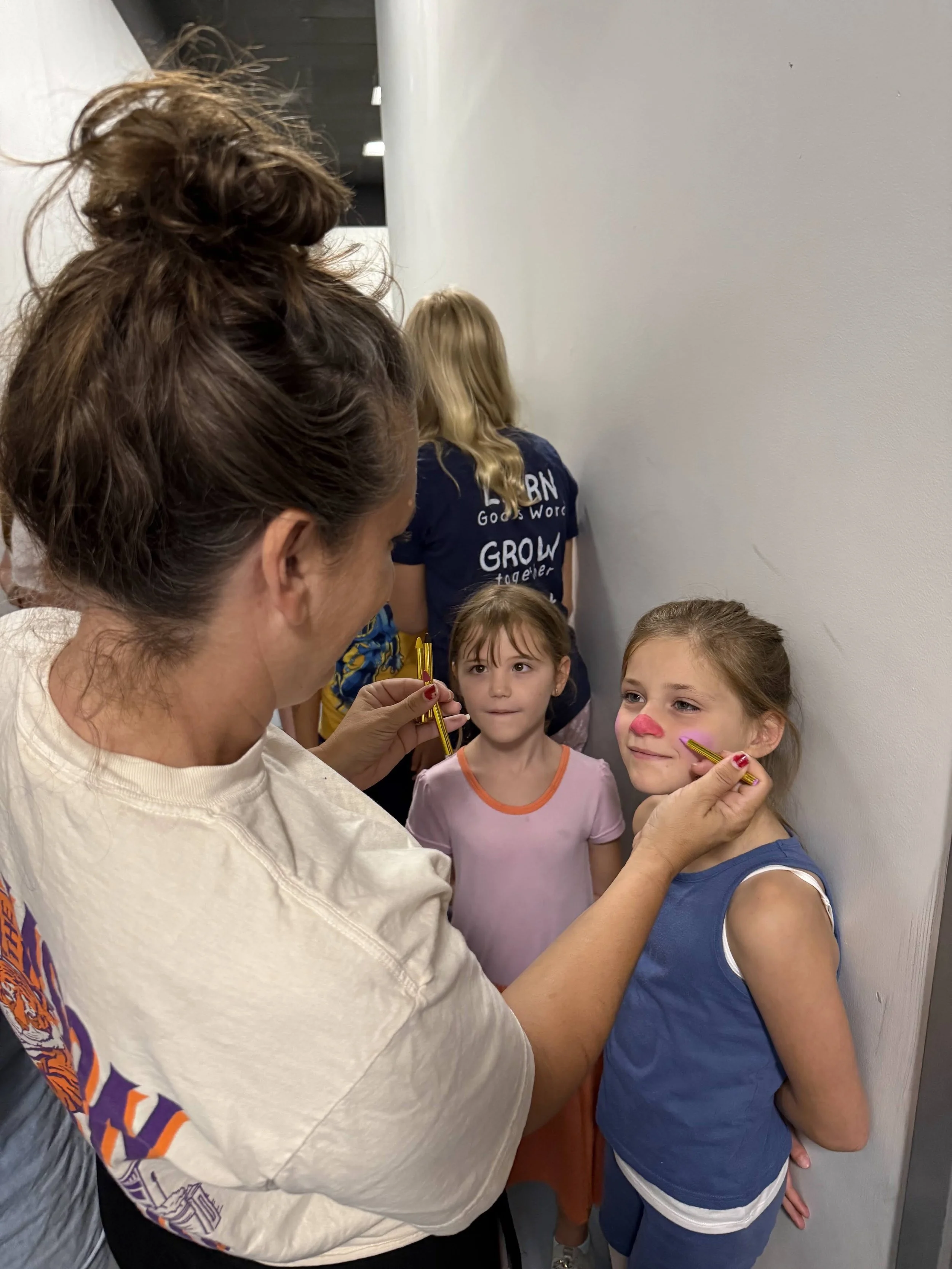 A adult volunteer applying pink face paint to a young performer backstage while another child looks on.