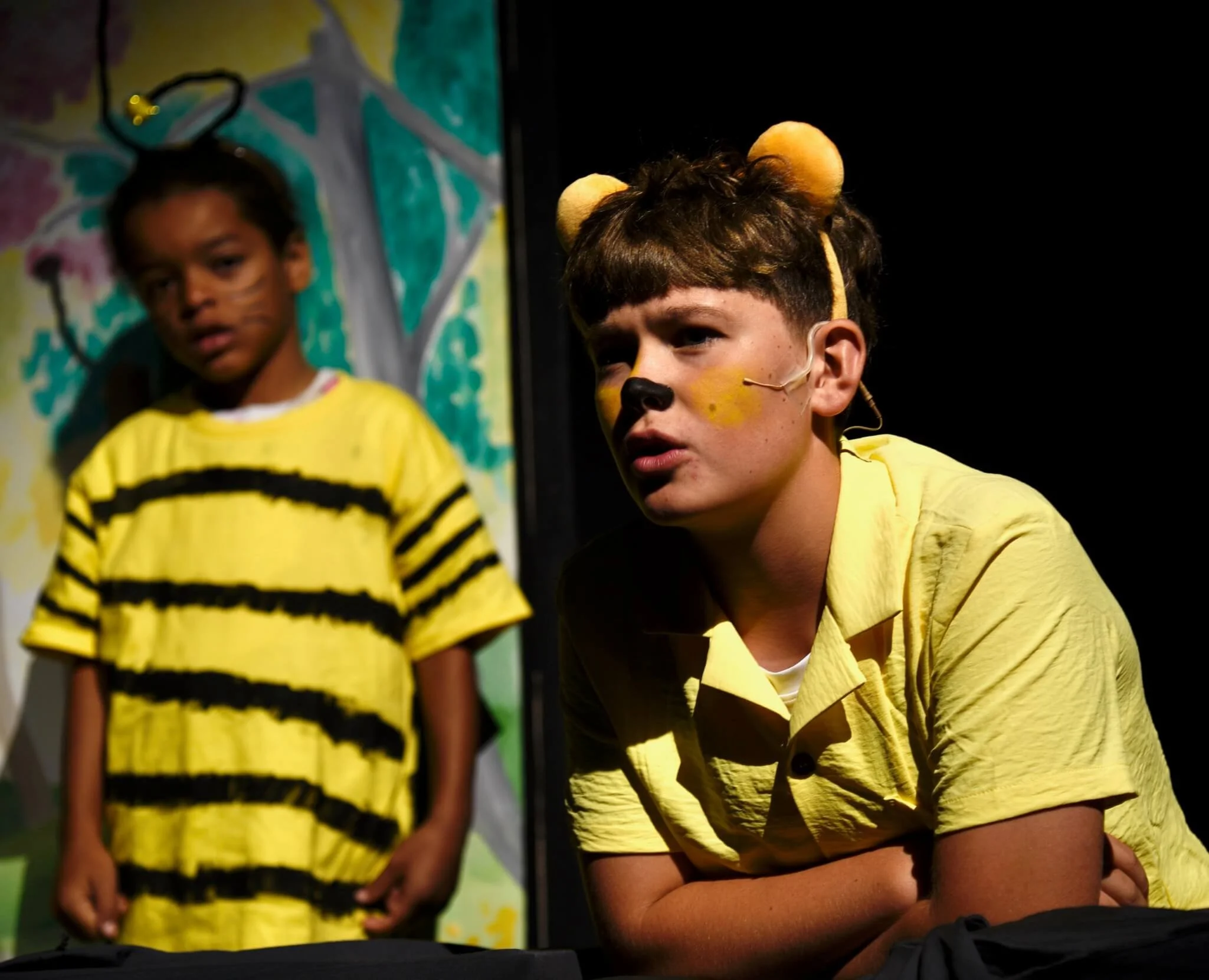 Close-up of a young actor on stage dressed as Winnie the Pooh in a yellow shirt, bear ears, and face paint, with another child in a bumblebee costume standing blurred in the background.