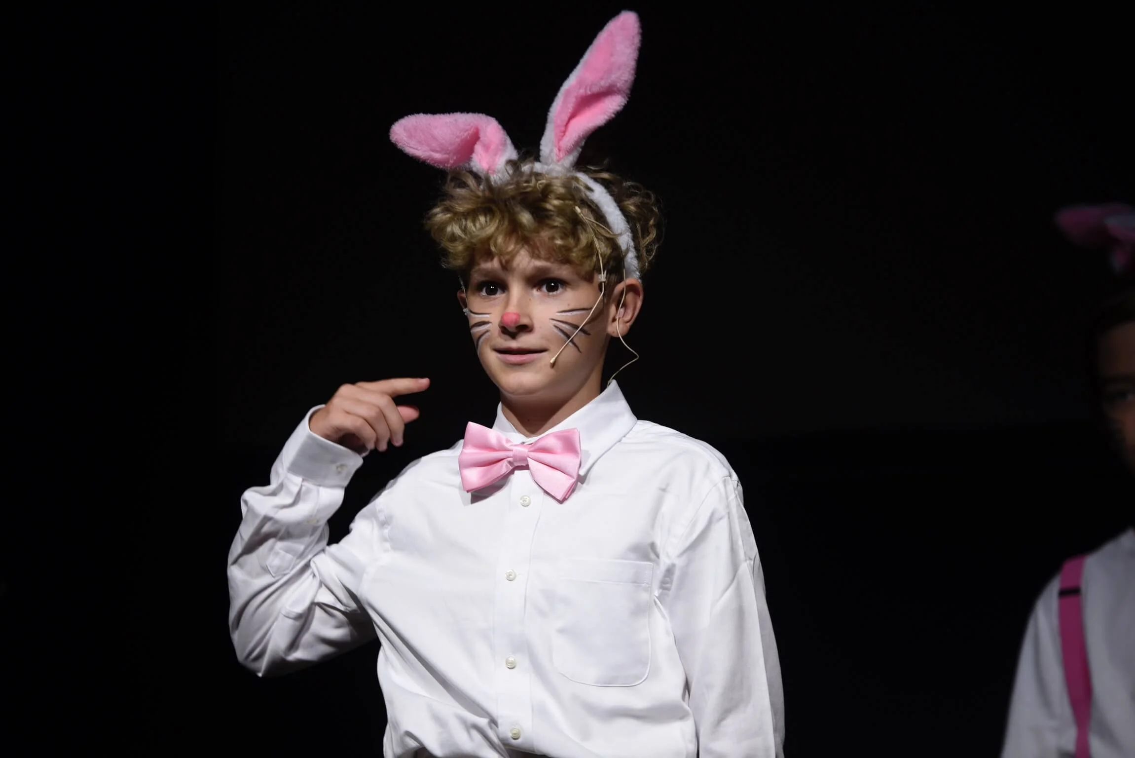 Close-up of a young actor wearing plush rabbit ears, a pink bow tie, and painted whiskers pointing to his head on stage.