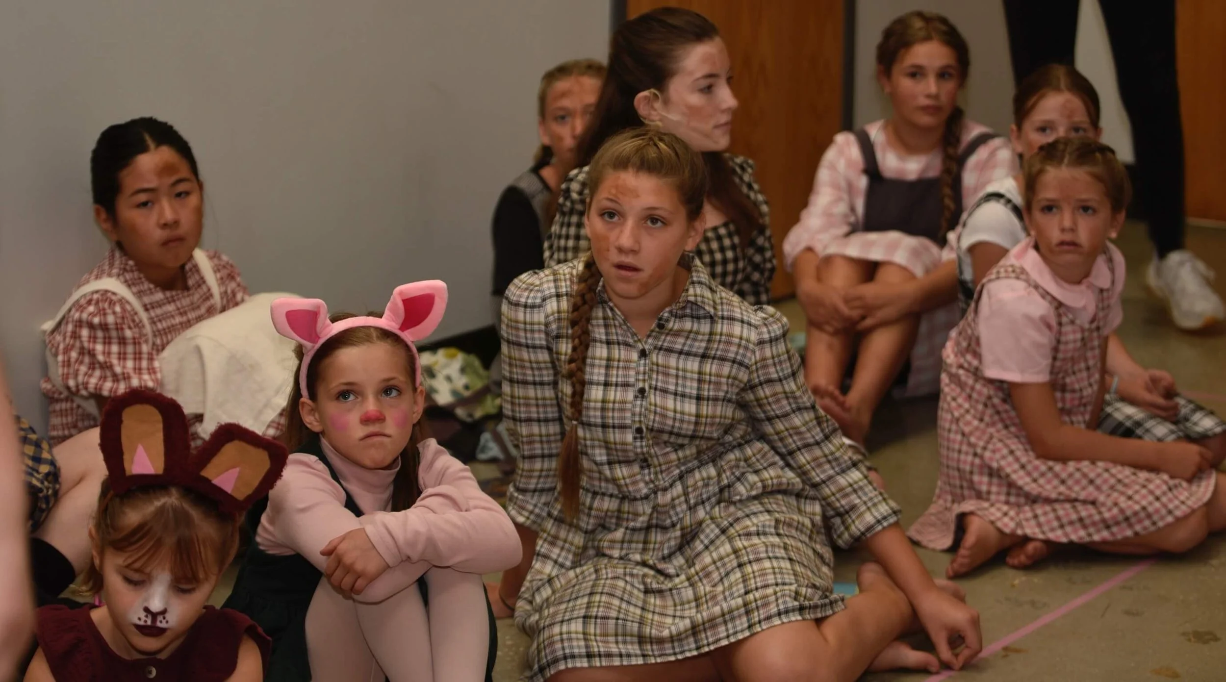 A group of young actors in costume sitting on the floor backstage, listening attentively.
