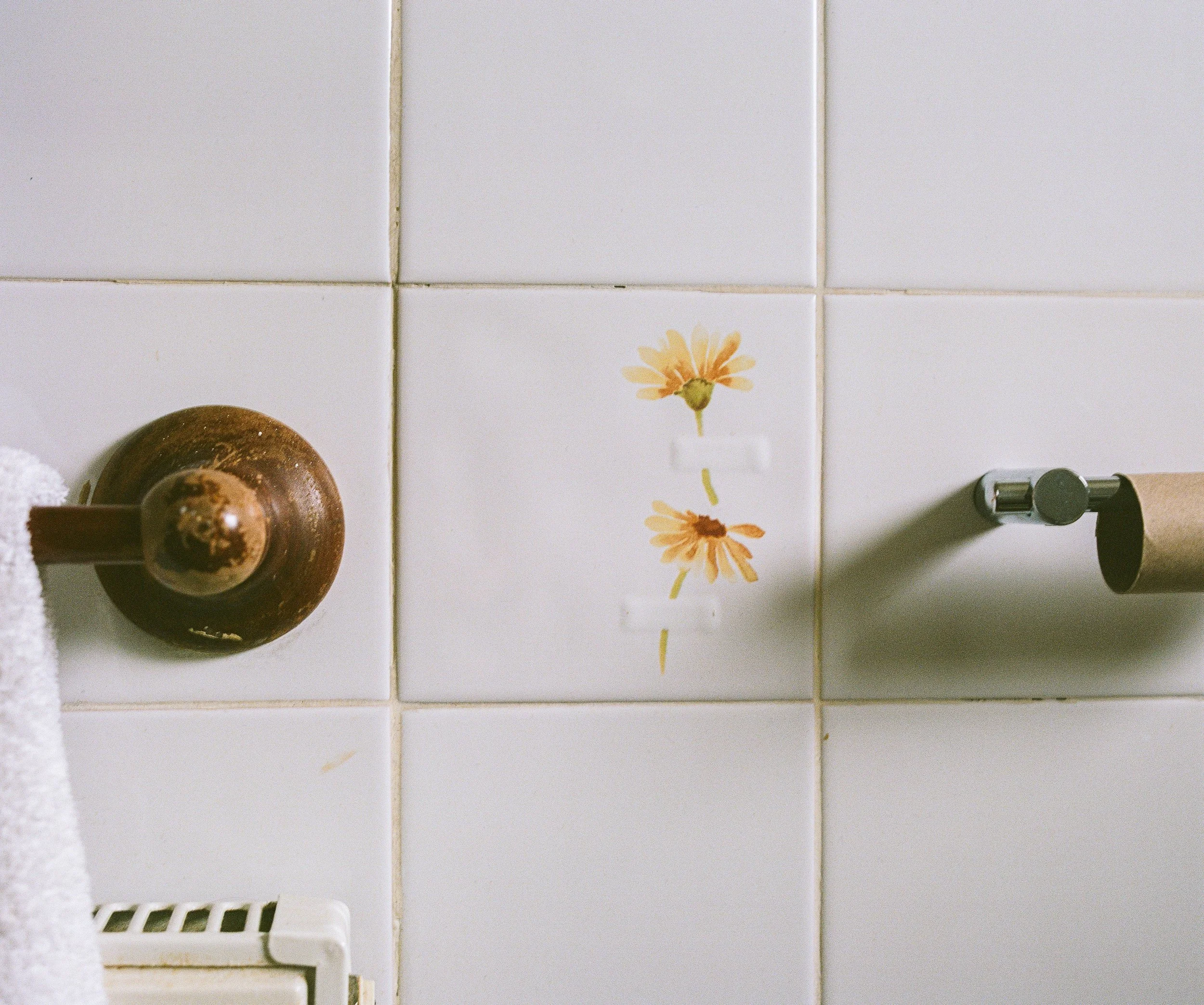 Close-up of a tiled bathroom wall with a floral sticker of two yellow daisies, a brown toilet paper holder on the left, a metal toilet paper roll holder on the right, and a white towel hanging on a radiator at the bottom.
