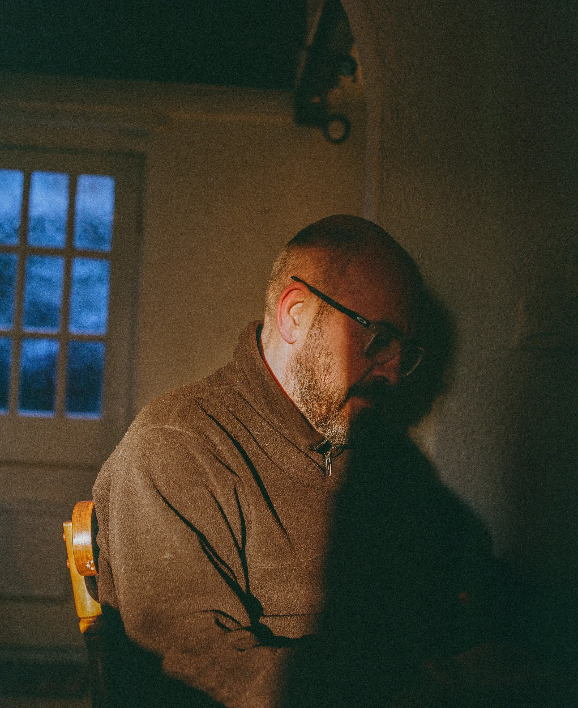 A man with glasses and a beard sitting indoors with a window in the background. He is wearing a brown zip-up sweater and appears to be focused on something not visible in the image.
