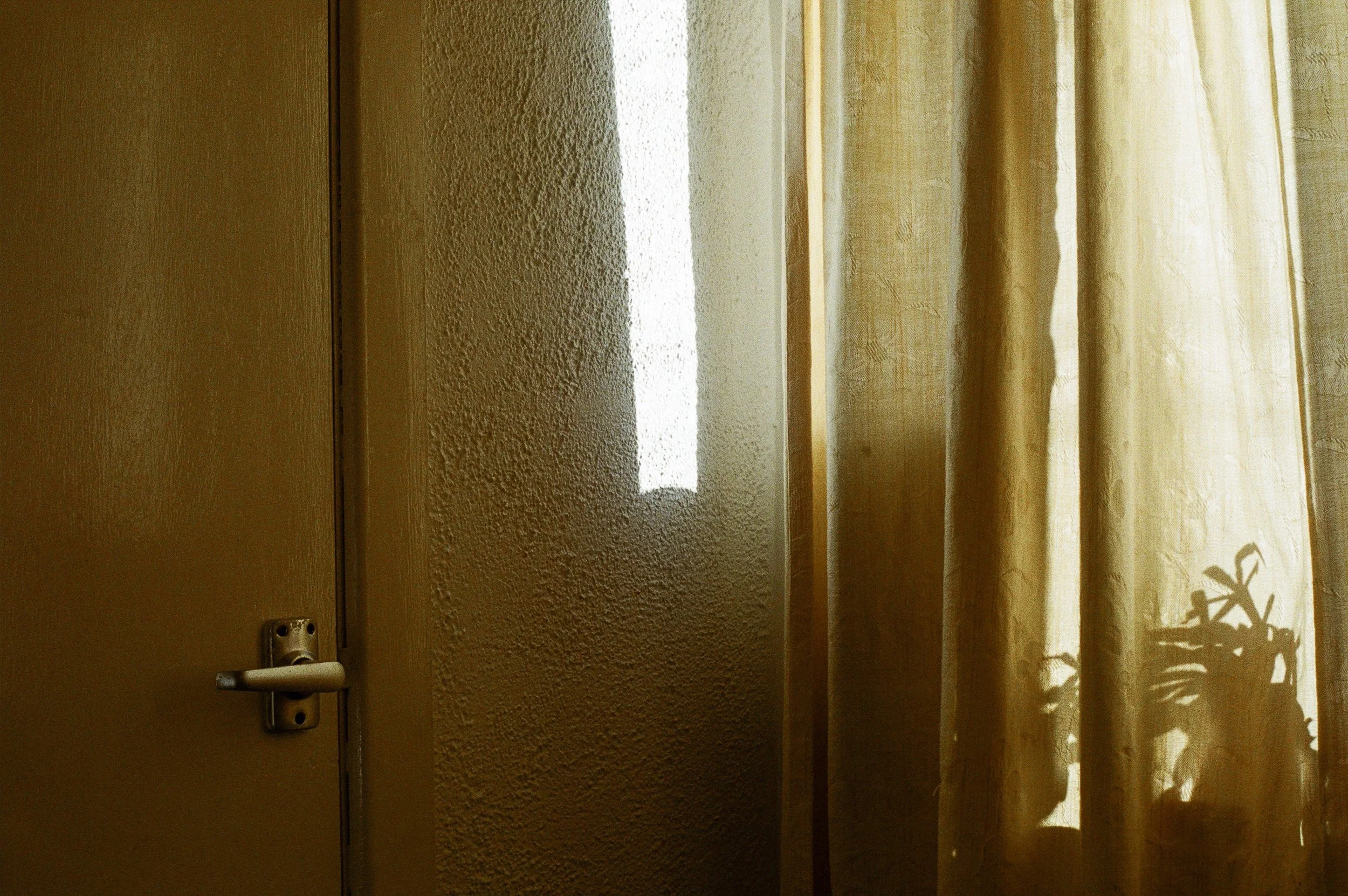 Sunlight shines through closed curtains onto a textured interior wall, with a door on the left side.