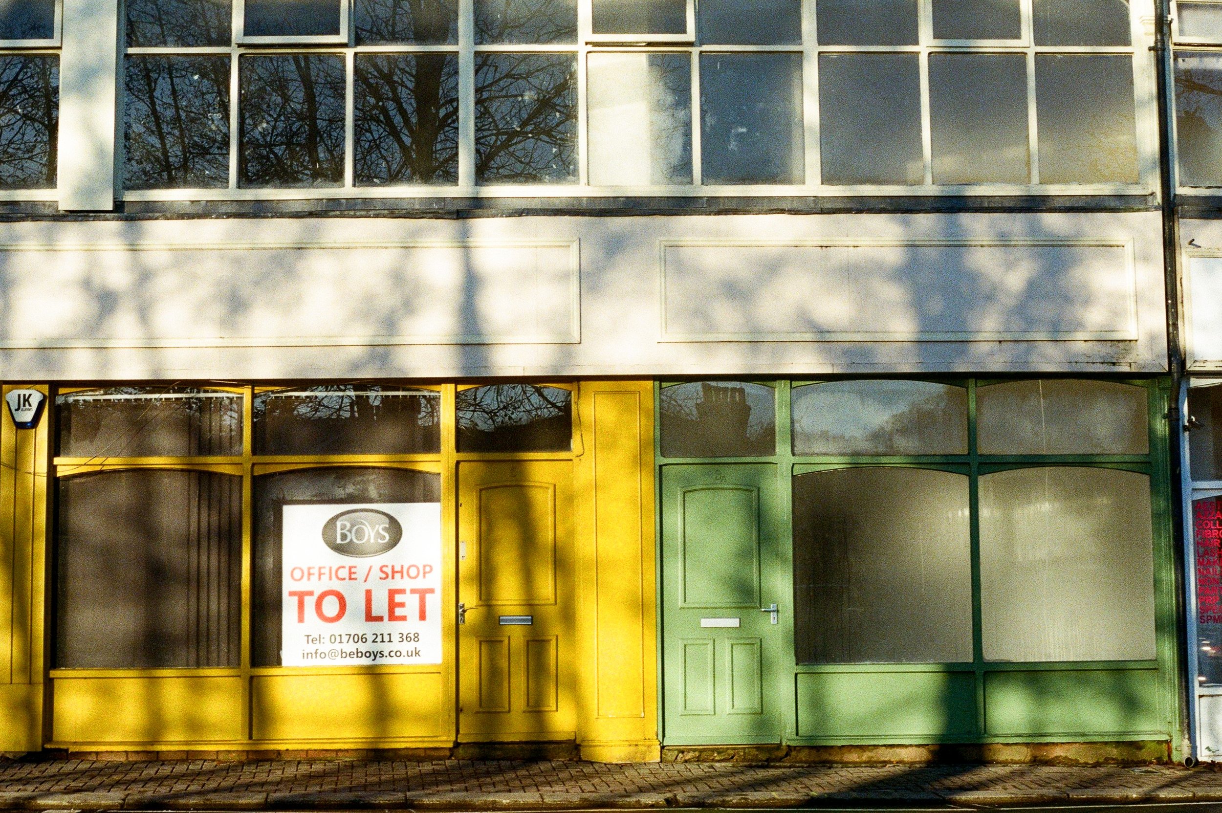Colorful storefront facade with yellow and green doors, a sign indicating the office/shop is available for rent, and trees reflected in the windows above.