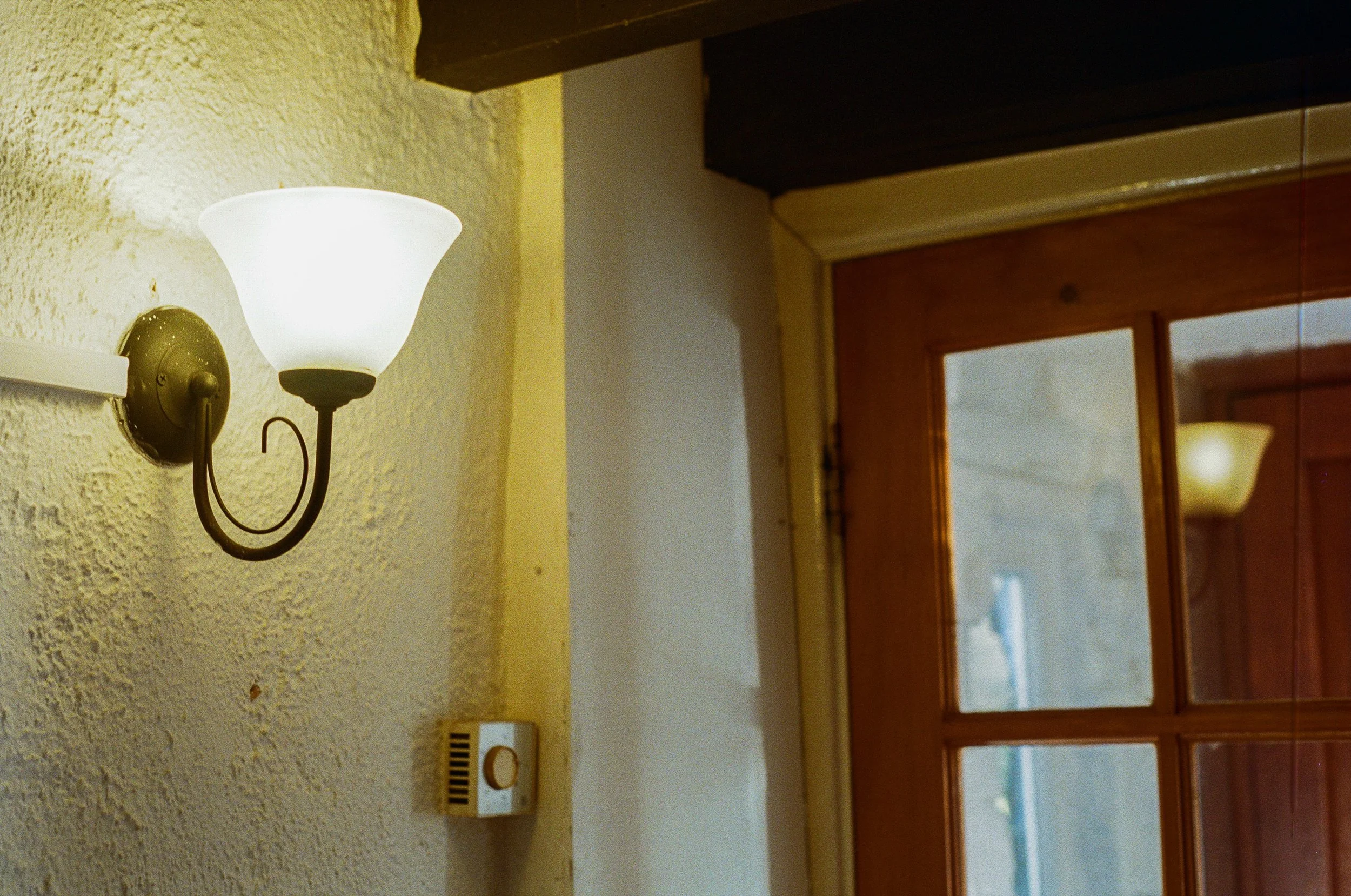 A wall-mounted light fixture with a white lampshade, next to a yellow textured wall and a wooden window frame.