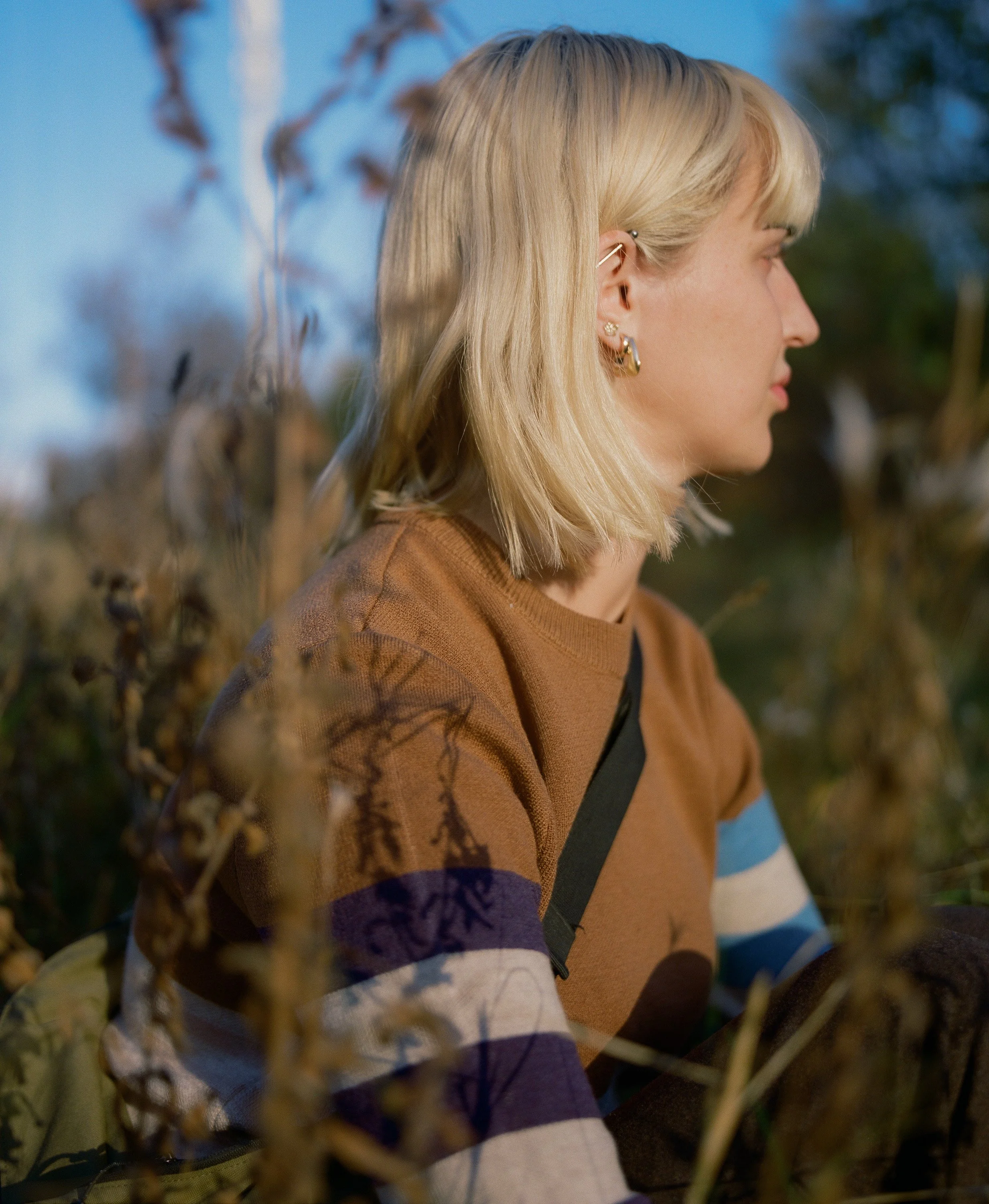 Profile of a young woman with blonde hair and multiple earrings, sitting outdoors among dried plants, wearing a striped sweater in brown, white, purple, and blue, with a patch of black strap visible.