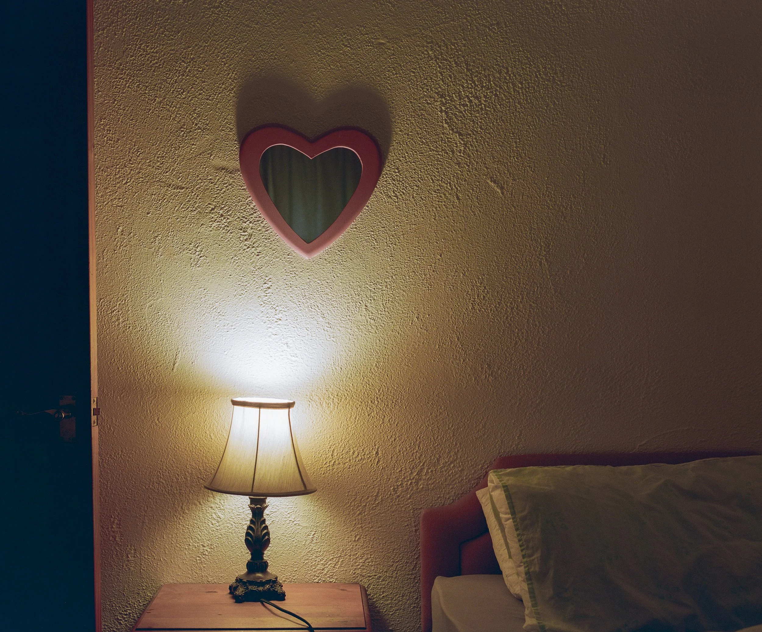 A bedroom corner with a wooden nightstand, a lit table lamp, a bed with a pillow, and a wall mirror shaped like a heart.