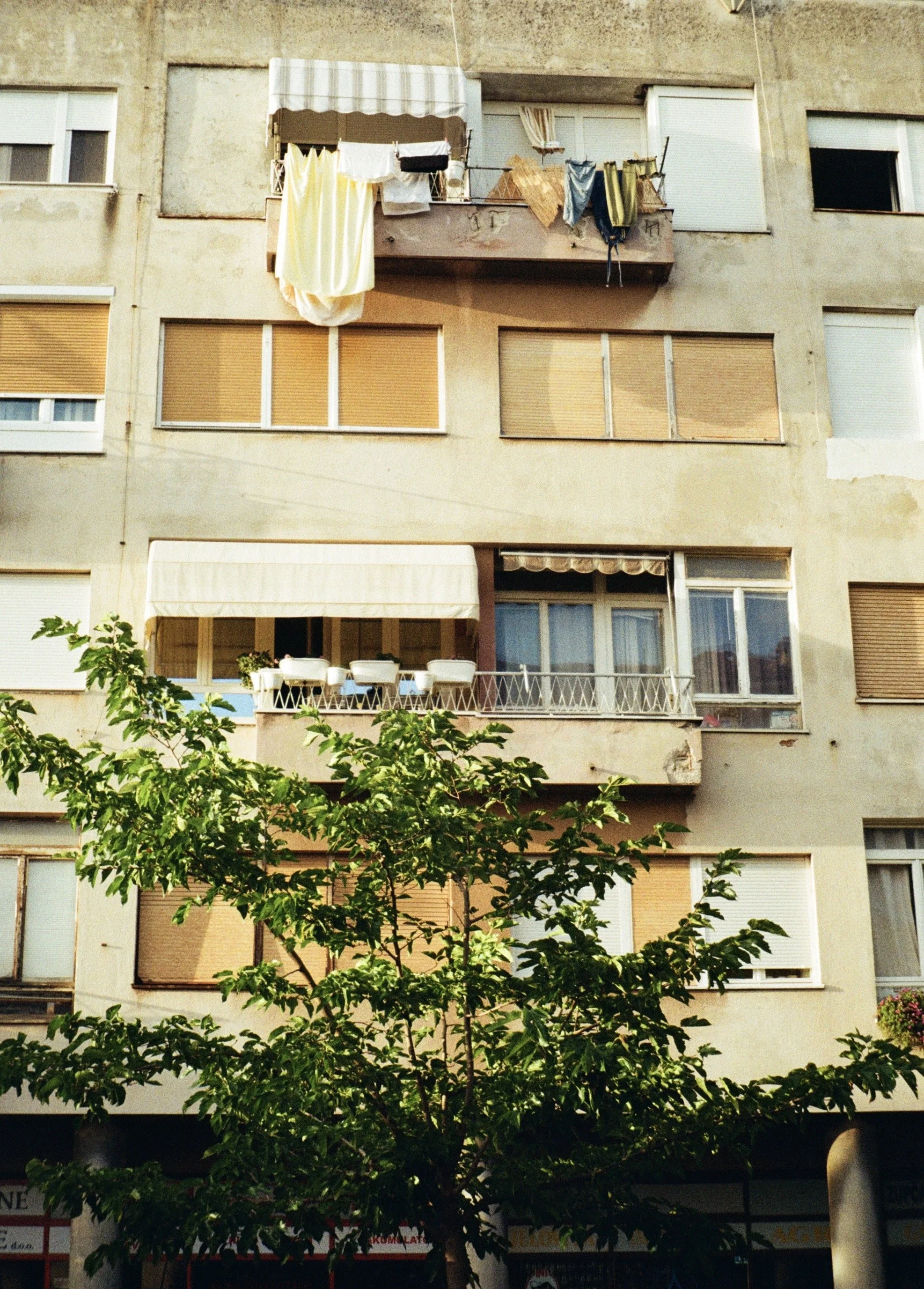 A multi-story residential building with some windows having blinds and some open, a balcony with chairs and plants, laundry hanging on the upper balcony, and a tree with green leaves in front.