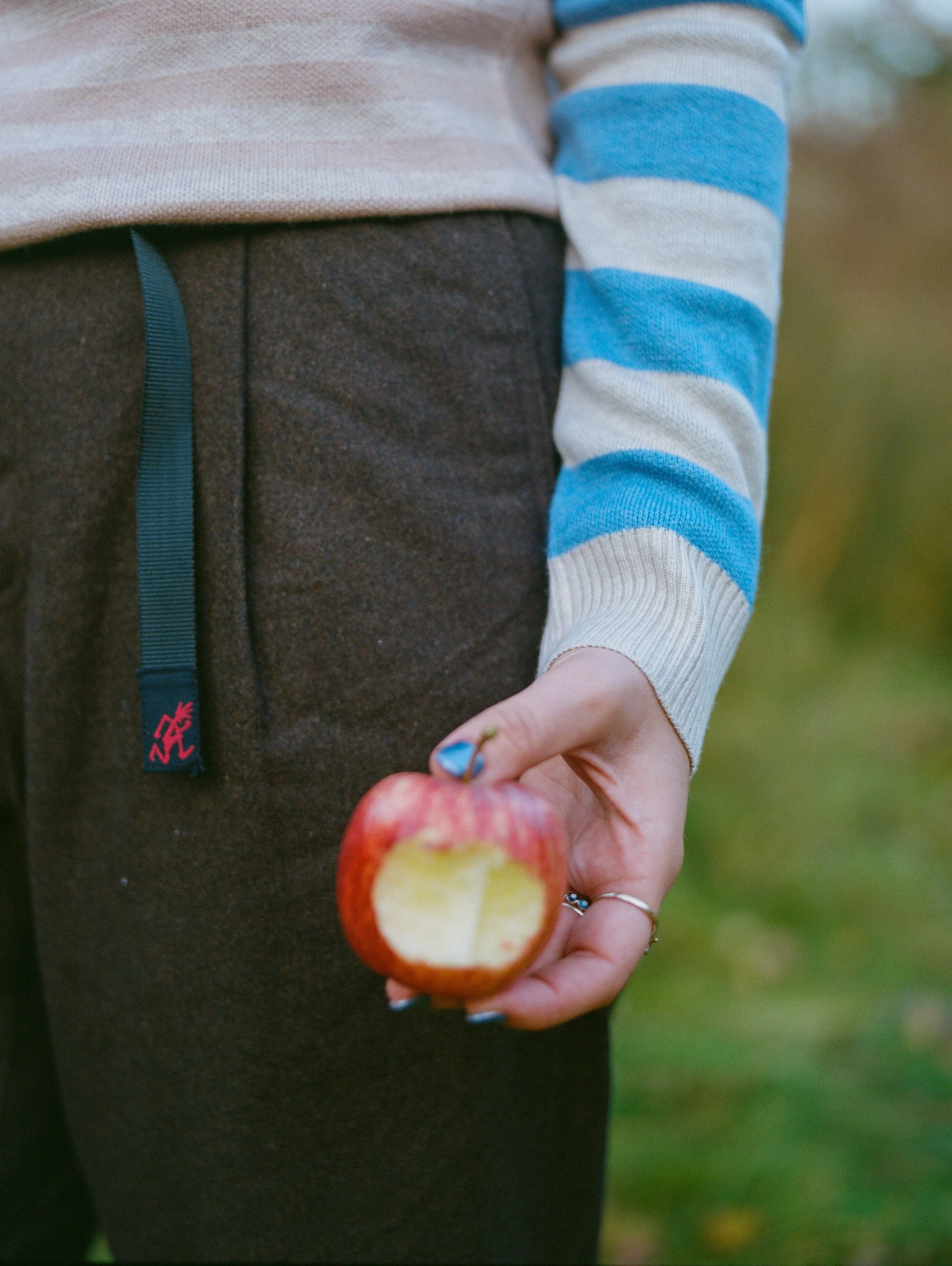 Person wearing dark pants and a beige and blue striped sweater holding a partially eaten red apple.