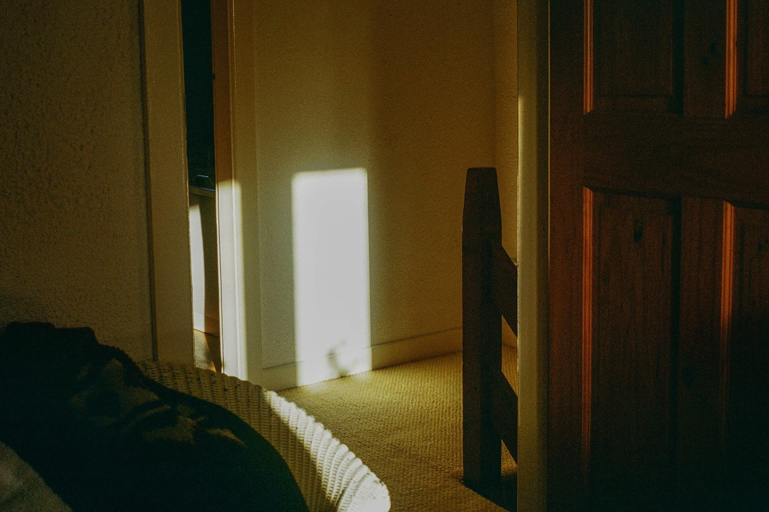 Interior of a room with sunlight streaming through an open door, casting bright rectangles on the wall and floor, with part of a wicker chair and a dark wooden cabinet visible.