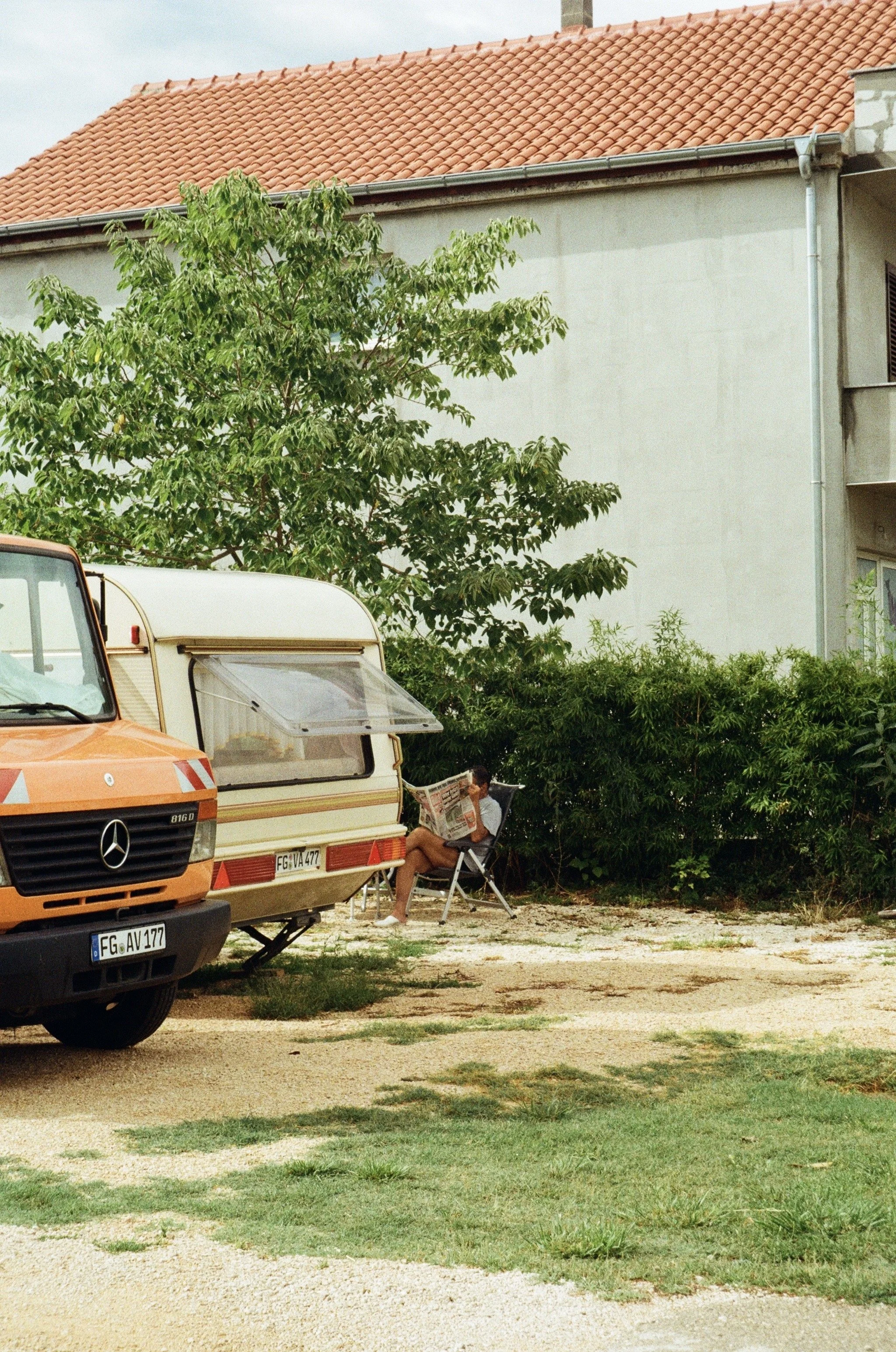 A woman sitting on a folding chair outside a caravan, reading a newspaper, with an orange vintage Mercedes-Benz van parked nearby. There's a small tree and some bushes in the background, and a building with a tiled roof.