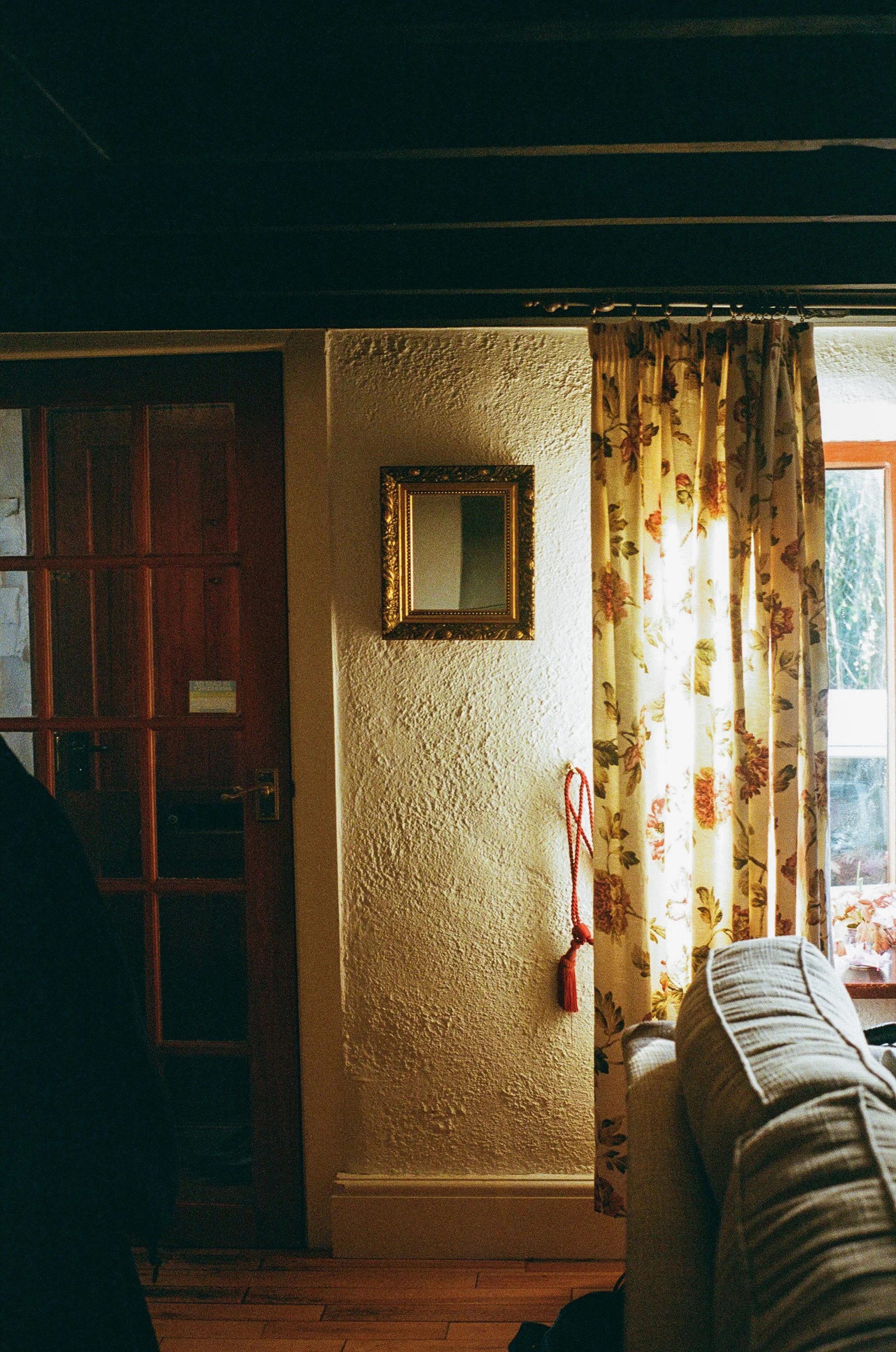 Interior view of a cozy living room corner with a textured wall, floral curtains, a mirror with a decorative gold frame, a sofa, and a window letting in natural light.