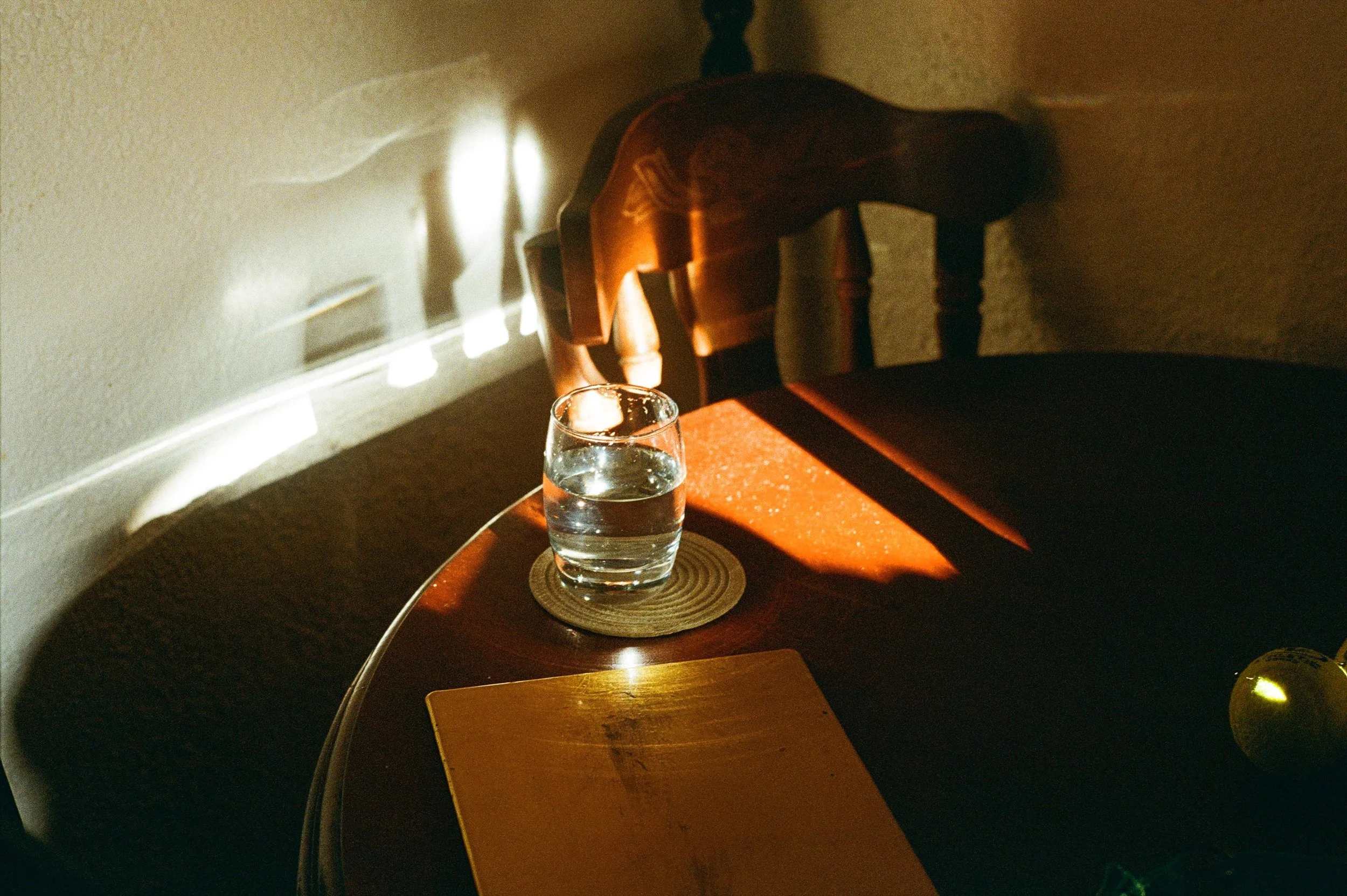 A glass of water on a round mat on a wooden table, with sunlight casting shadows on the table and wall behind, and a book or notebook partially visible.