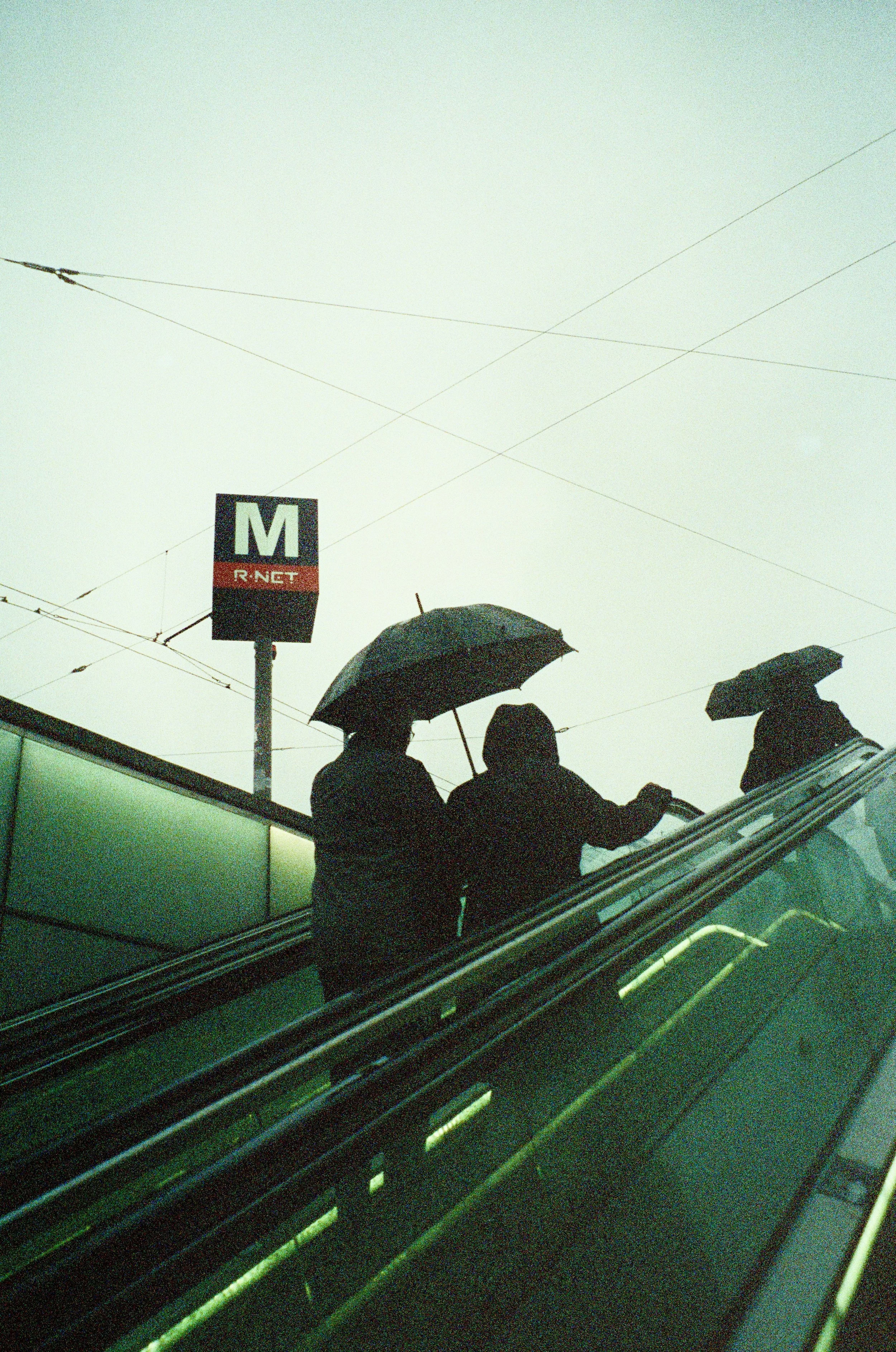 Silhouettes of three people with umbrellas on an escalator against a gray sky, with an M subway sign in the background.