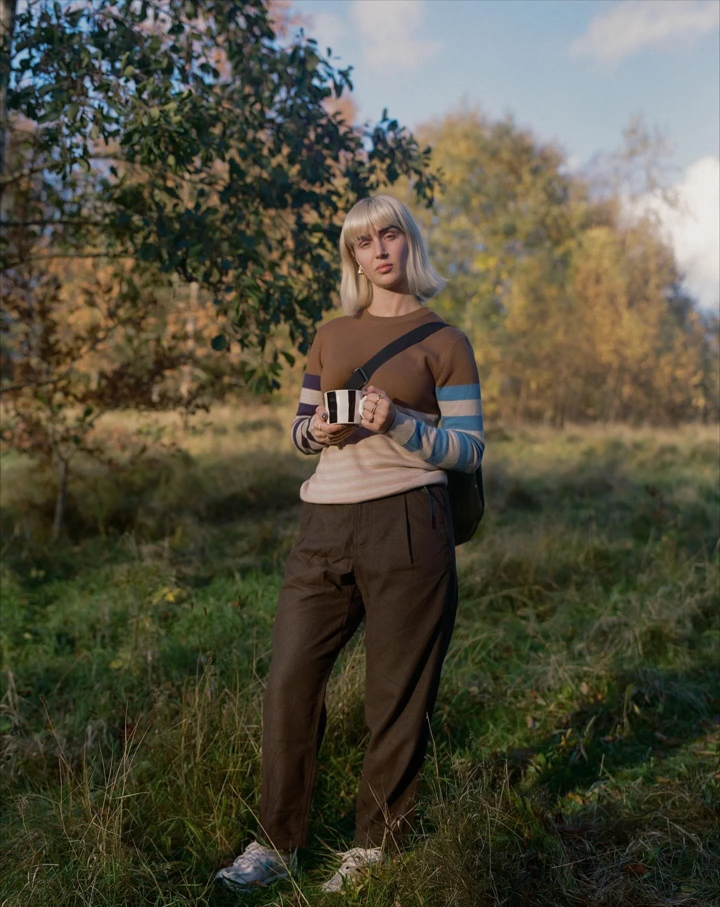 A young woman with blonde hair in a bob, wearing a striped brown and blue sweater, brown pants, and white sneakers, stands outdoors in a grassy area with trees and a cloudy sky in the background, holding a black-and-white striped mug.