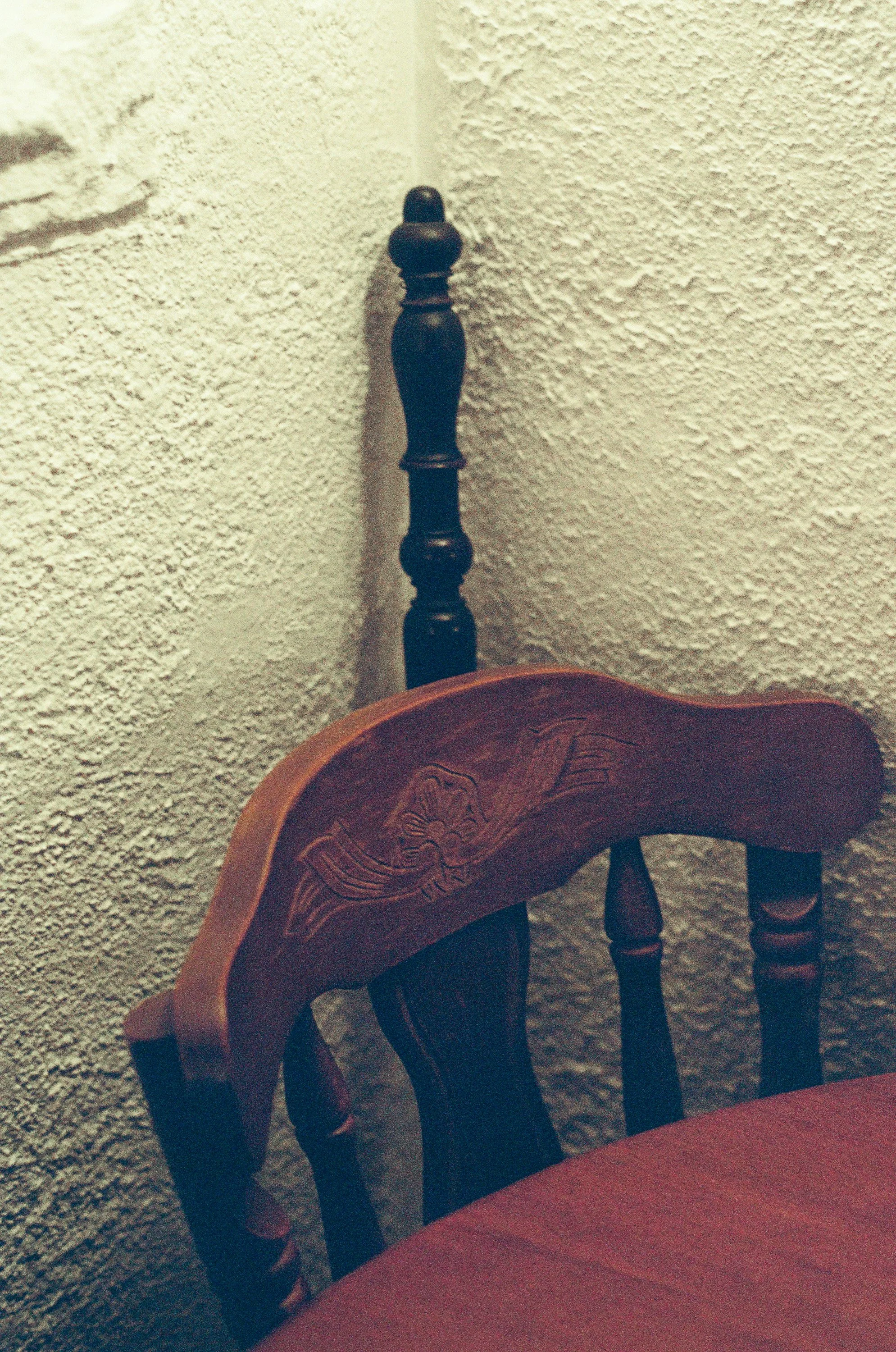Close-up of a dark wooden dining chair with a carved floral design on the backrest against a textured yellow wall.