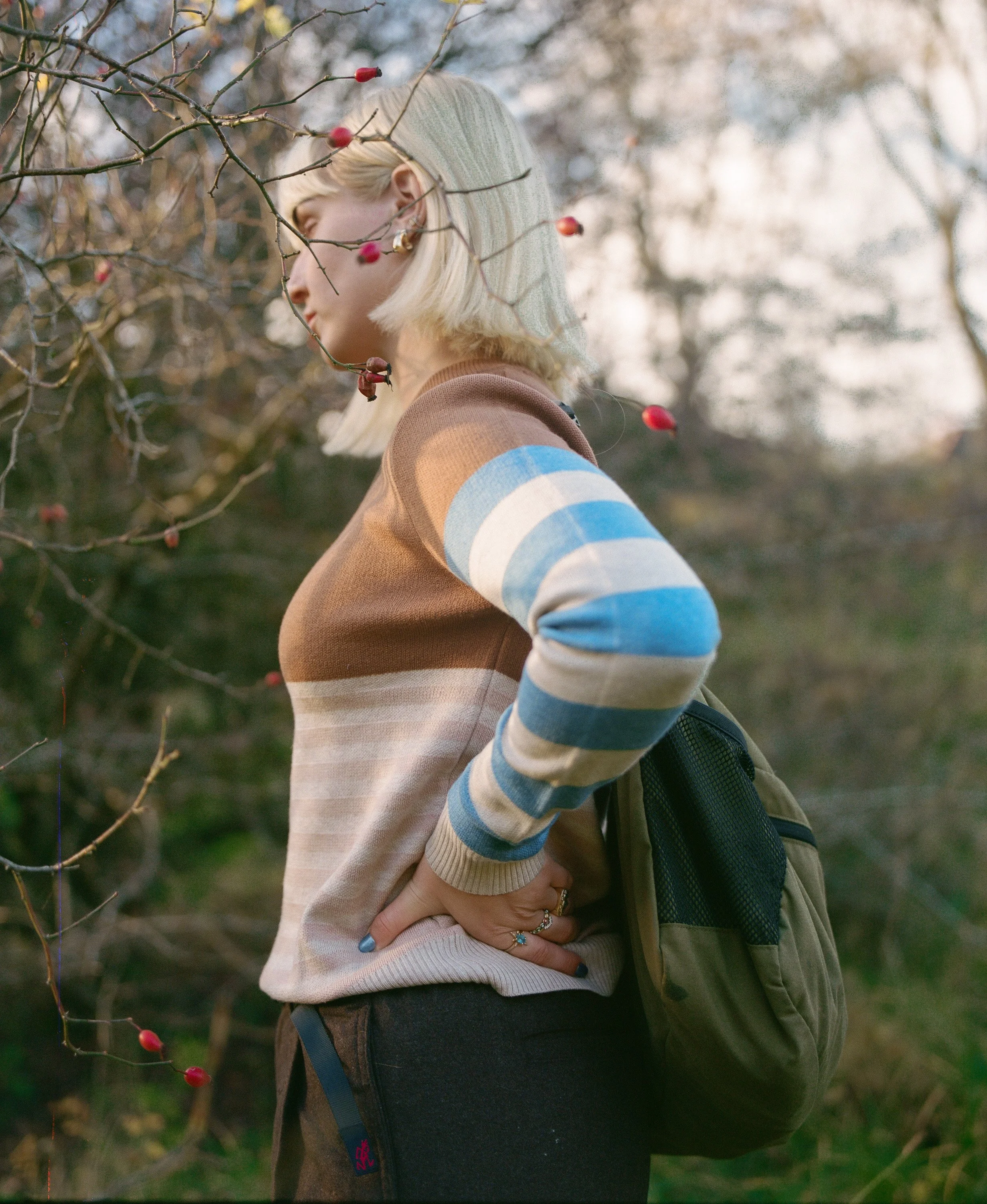 A woman with blonde hair wearing a striped sweater and a beanie, standing outdoors near a tree with red berries, with her hand on her hip and a backpack on her shoulder.