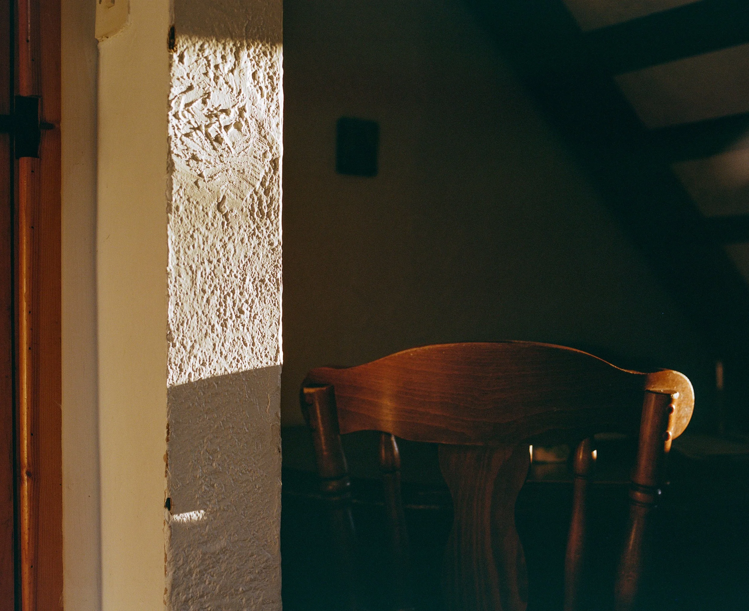 Sunlight illuminates a textured wall and a wooden chair in a dim room.