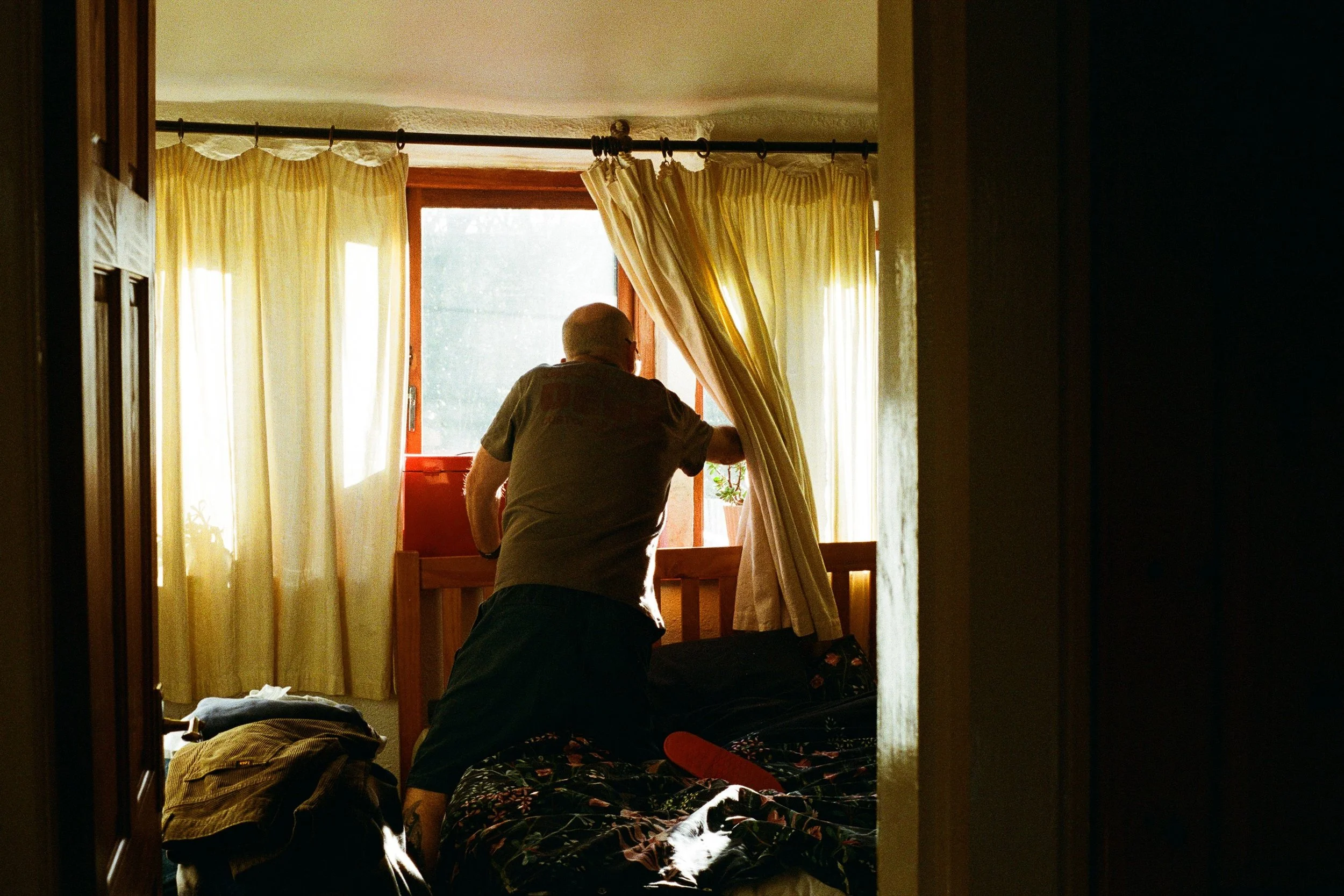 A man standing by a window with yellow curtains, reaching out to touch a potted plant in a bedroom.
