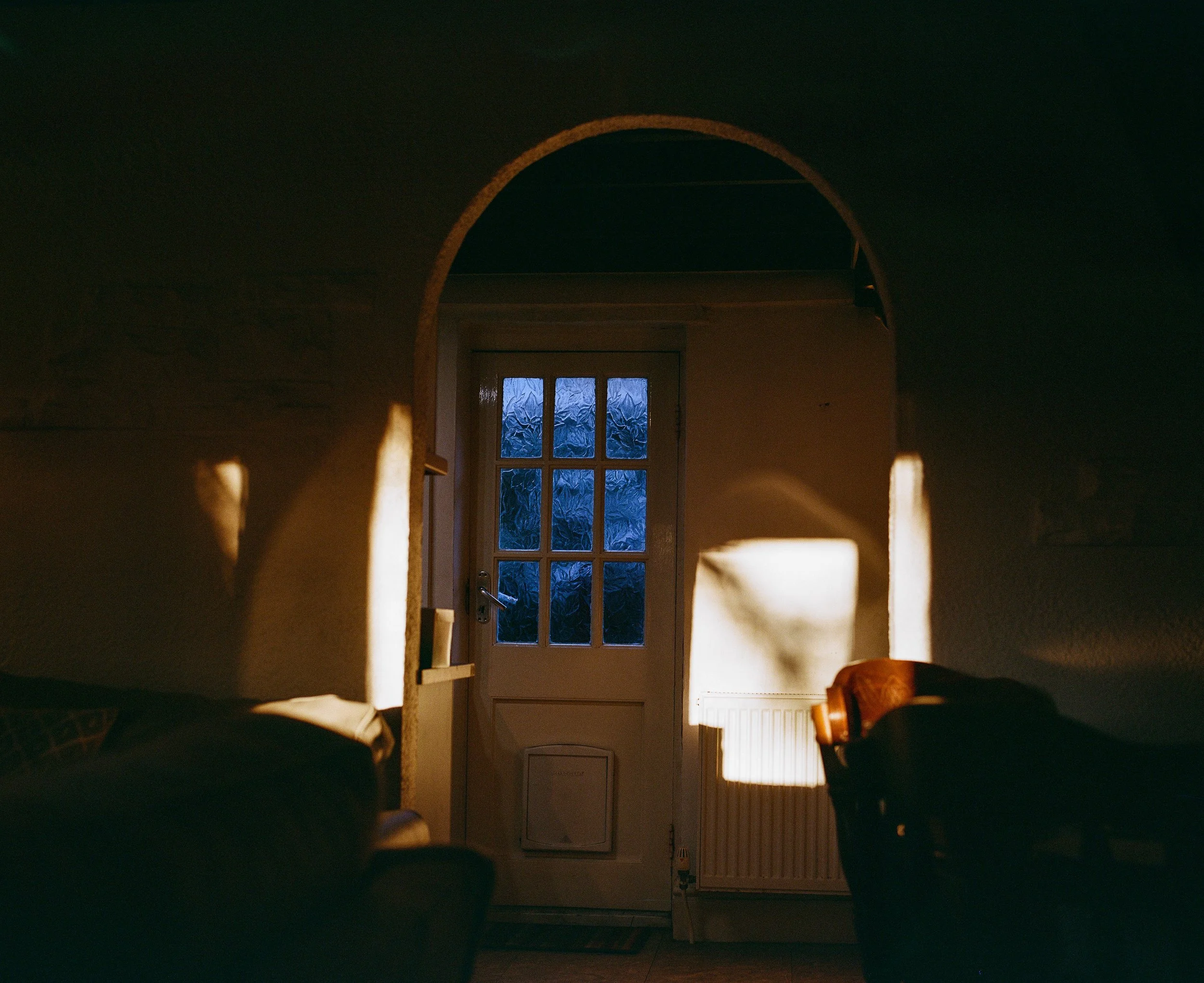 Sunlight casts shadows on a white door with glass panels, seen through an arched doorway in a dimly lit room.