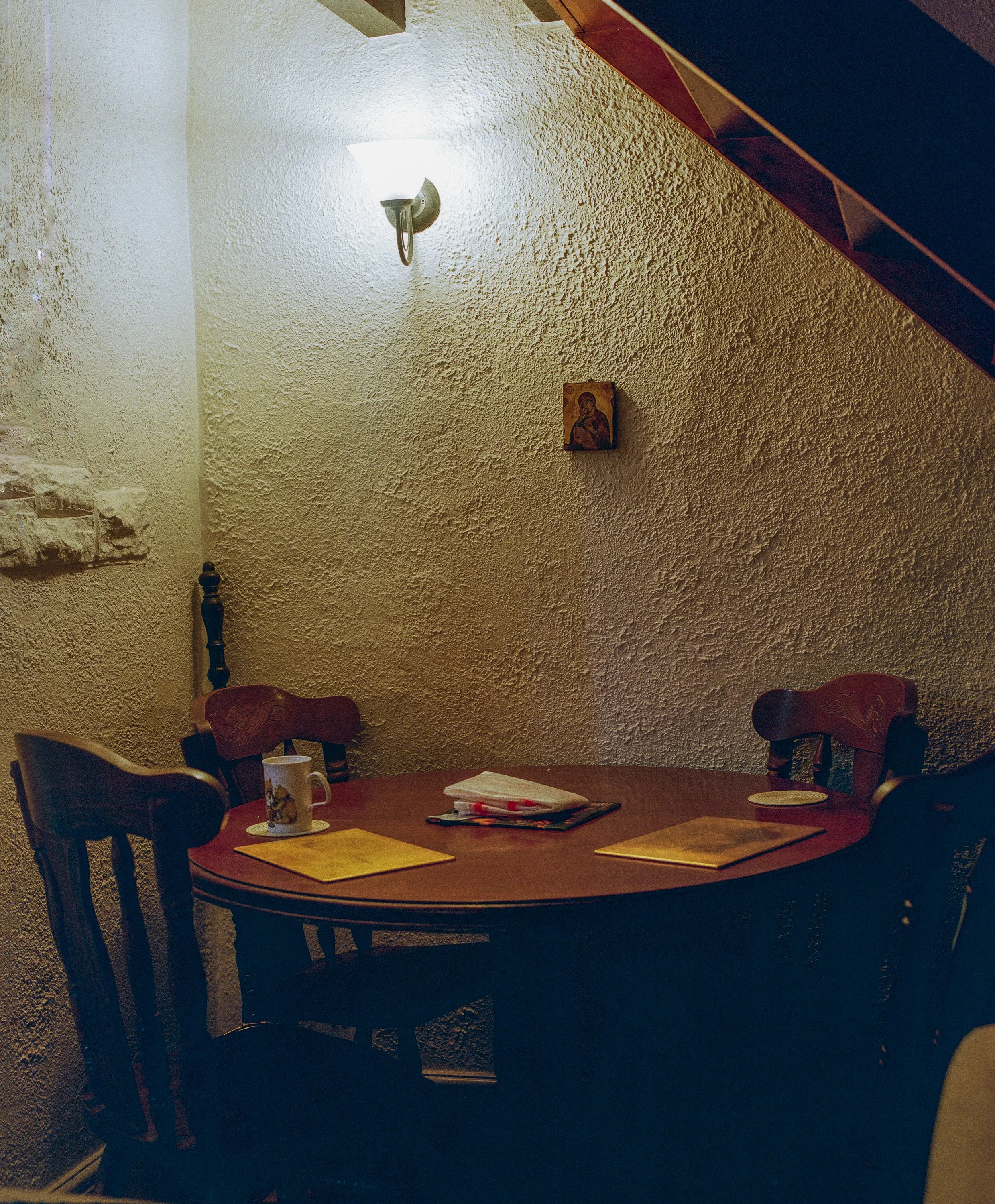 A cozy dining nook with a round wooden table set with yellow placemats, a mug, and napkins, surrounded by four wooden chairs against textured beige walls with a small religious icon and wall-mounted light.