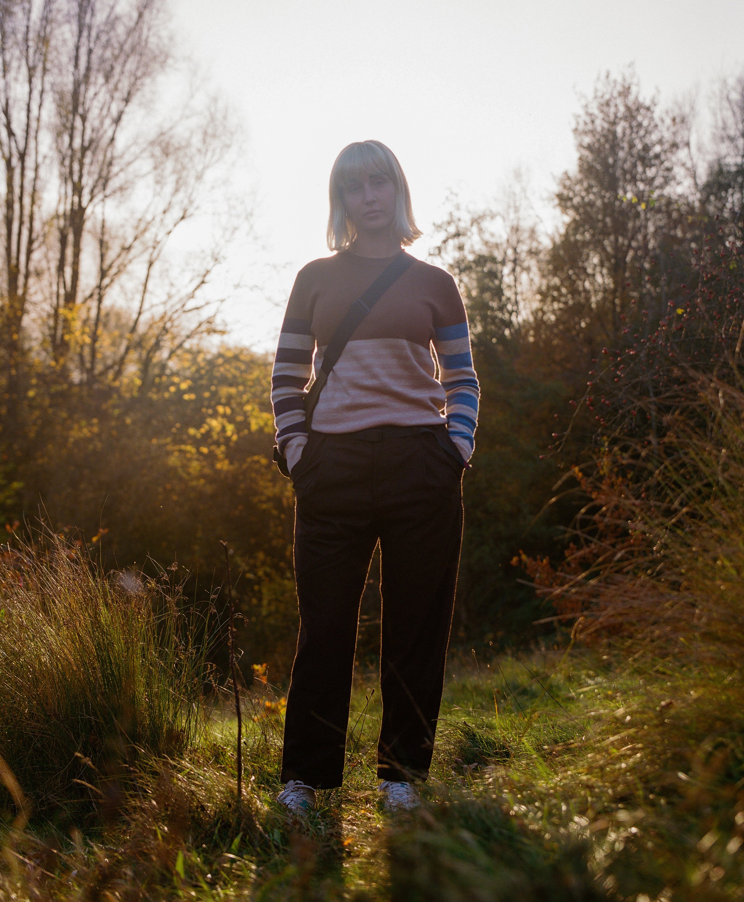 A woman standing outdoors in nature during sunset, wearing a striped sweater and black pants, with hands in her pockets.