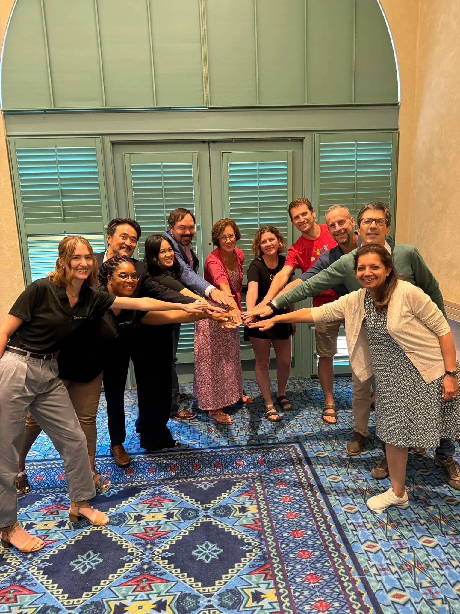 A diverse group of eleven people standing in a semi-circle with their hands stacked together in the center, smiling at the camera in a room with blue shutters and a patterned carpet.