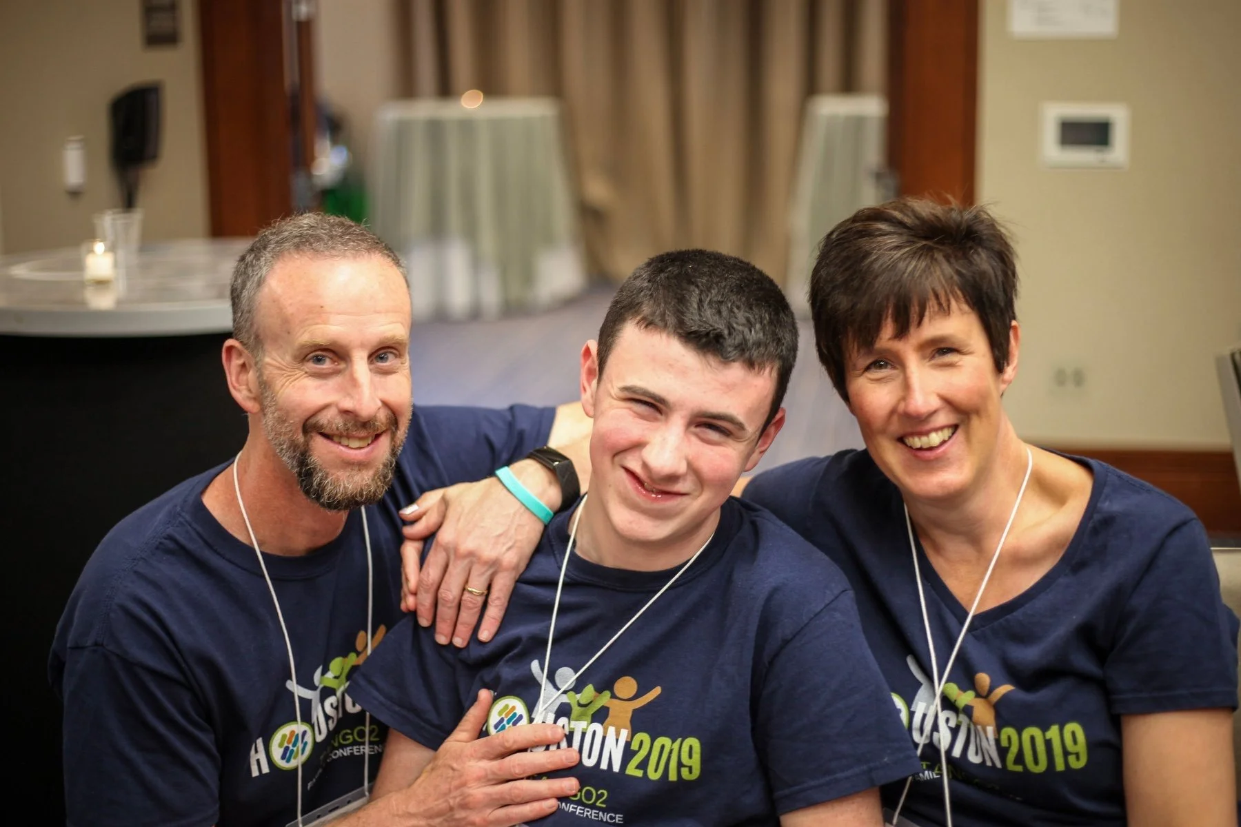 Three smiling individuals, two adults and one young person, wearing dark blue conference T-shirts with 'Houston 2019' printed on them, sitting close together with arms around each other in an indoor setting.