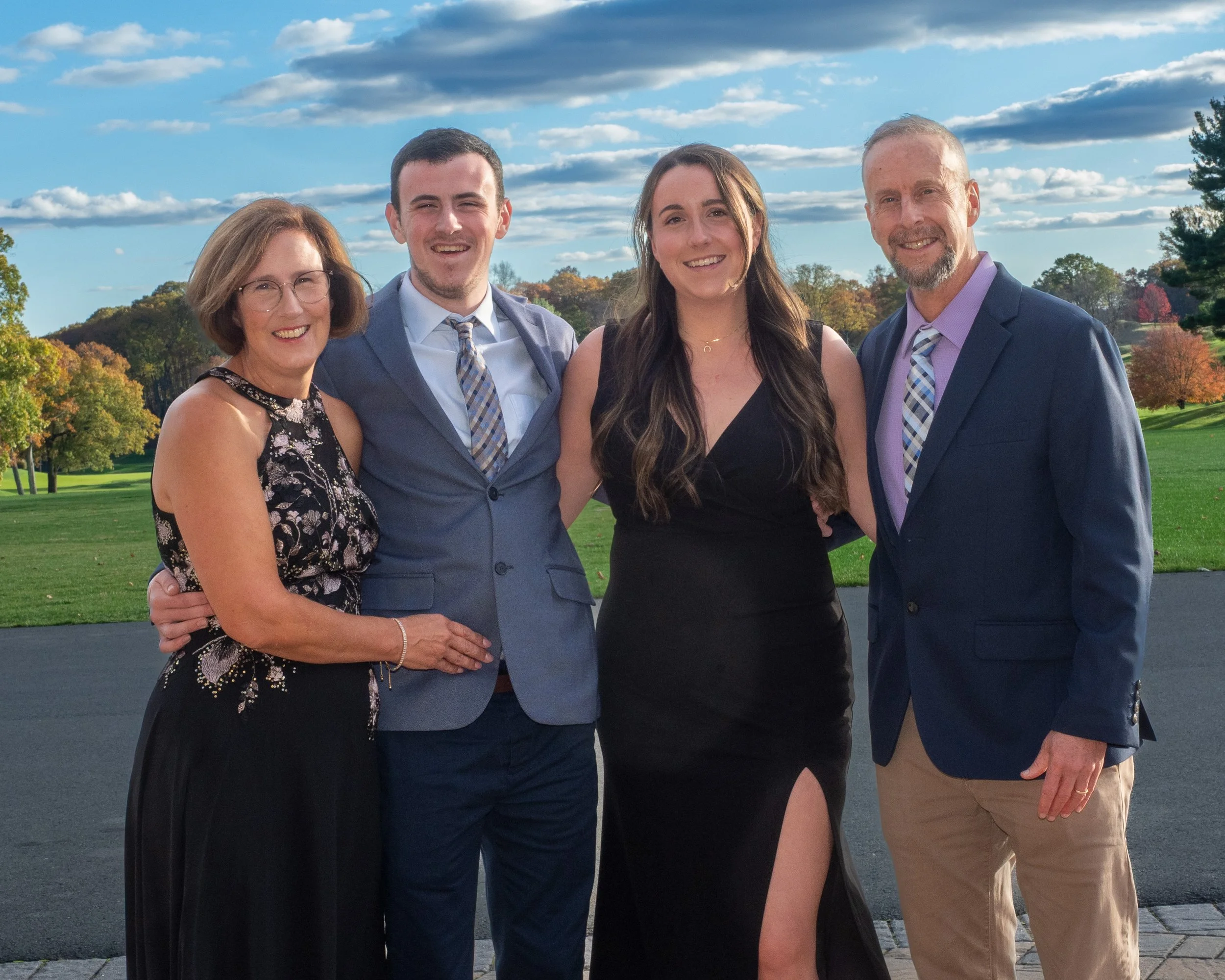 A family of four standing outdoors on a paved area with a grassy park and trees displaying fall foliage in the background, all smiling at the camera.
