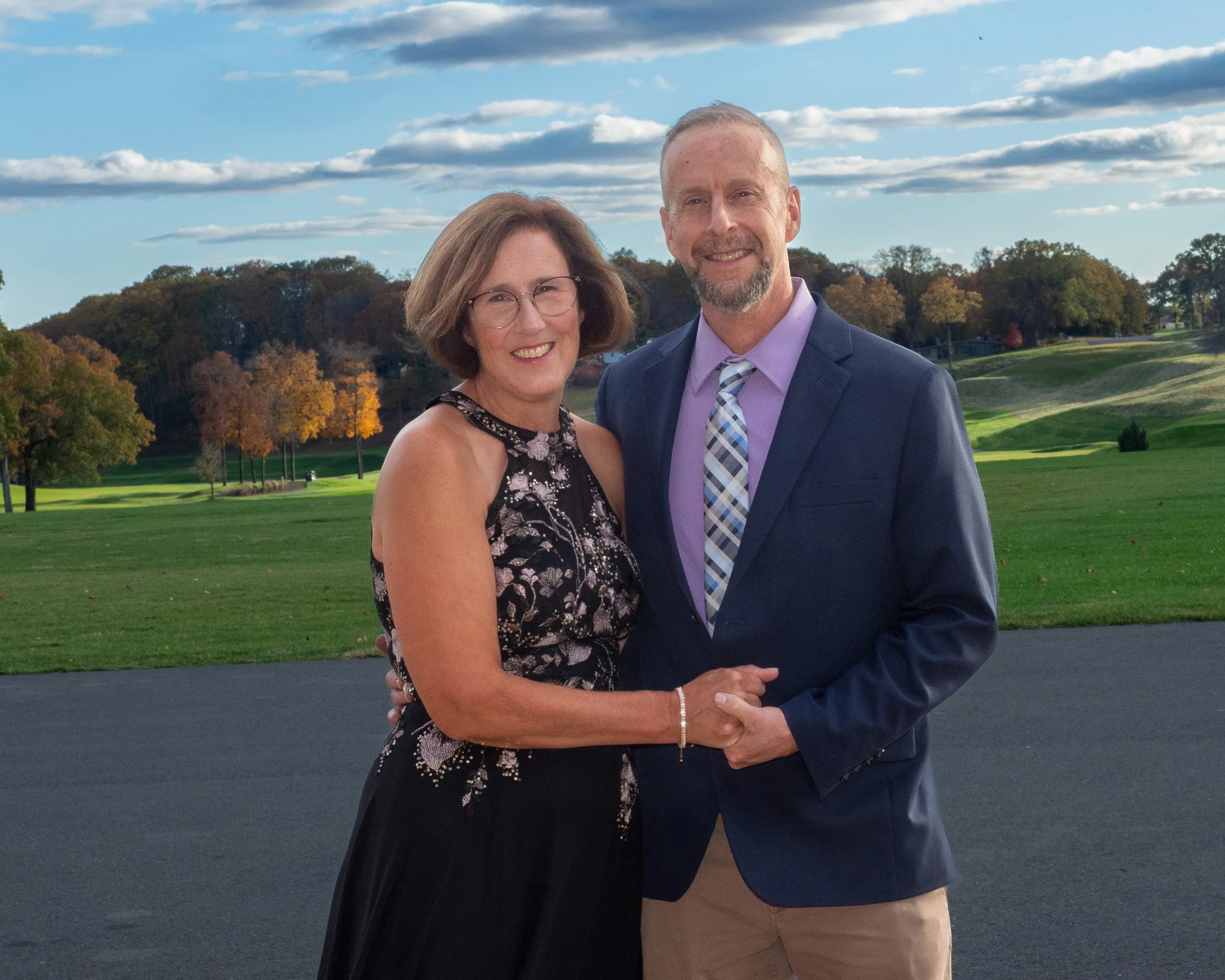 A smiling middle-aged woman and man in formal attire standing outdoors on a sunny day at a park or golf course, holding hands and facing the camera.